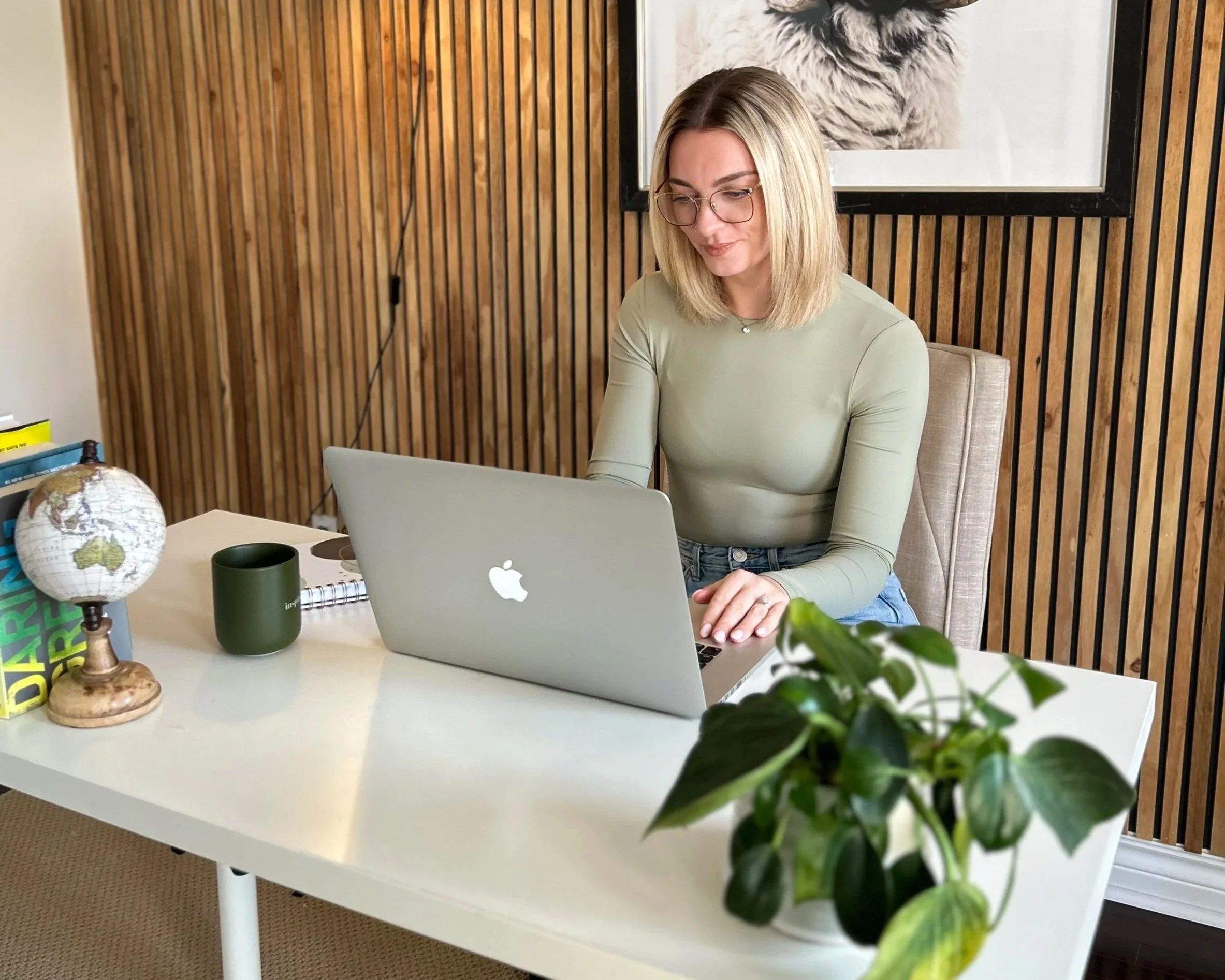 A woman with blonde hair and glasses sitting at a white desk, working on a silver MacBook in a room with wooden paneling on the wall. The desk has a globe, a green mug, a notebook, and books. A potted plant with green leaves is in the foreground, and a framed art piece featuring an animal is on the wall behind her.