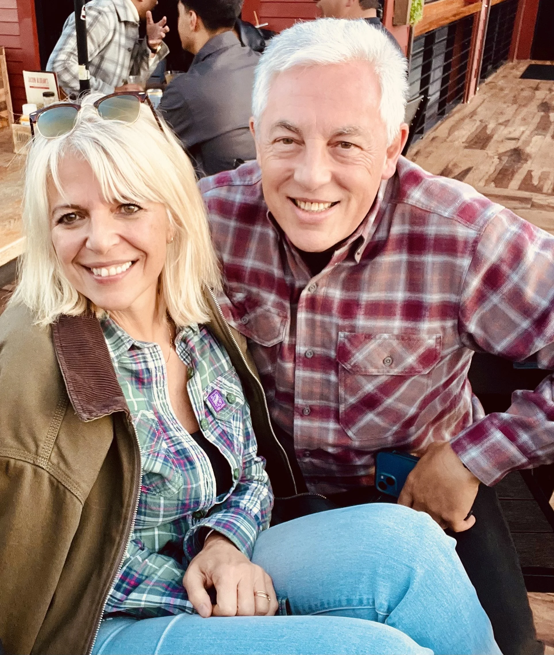 Smiling woman with blonde hair wearing sunglasses on her head, plaid shirt, and brown jacket sitting next to a smiling man with grey hair in a red plaid shirt at a wooden table restaurant with other people in the background.