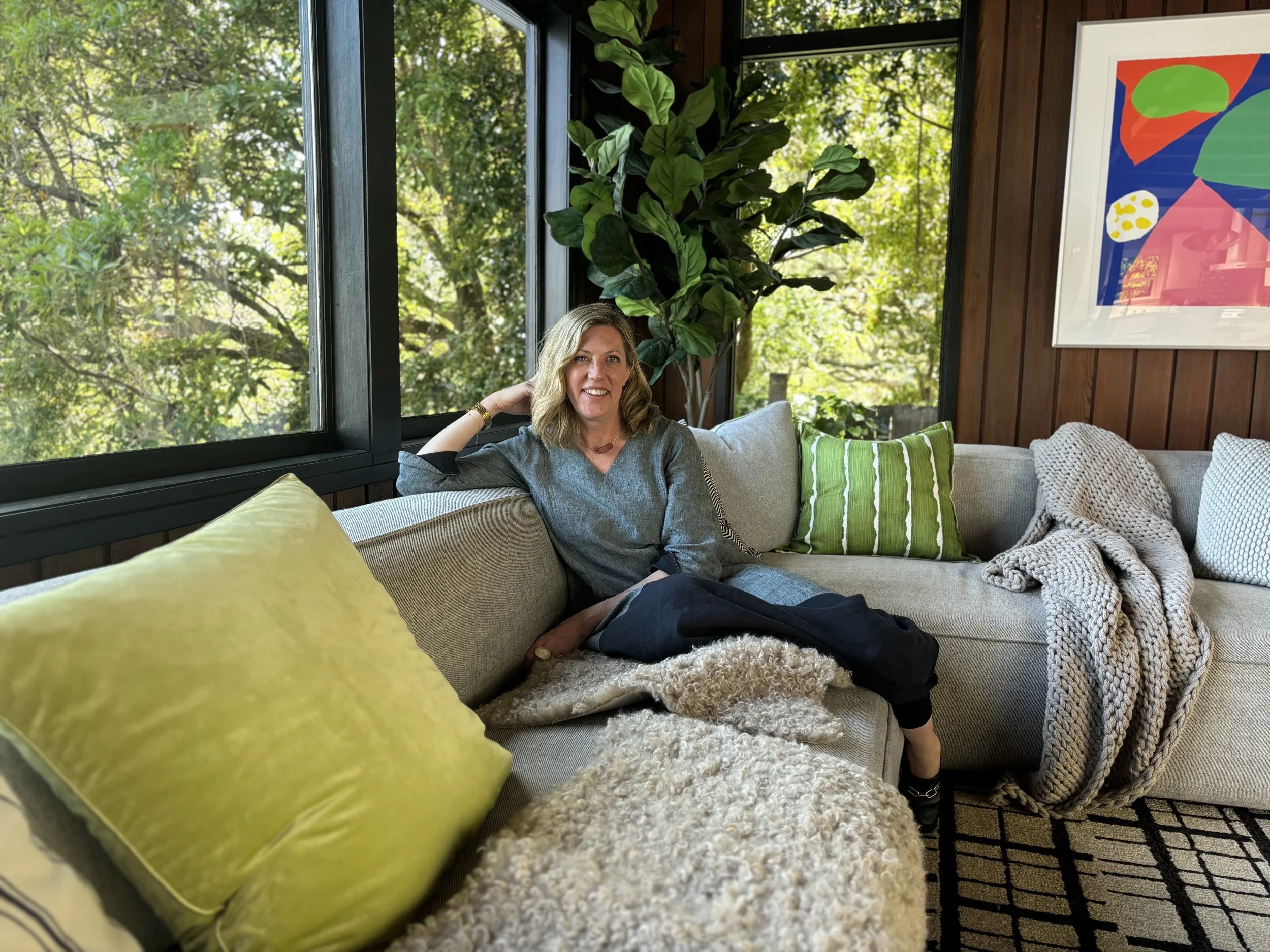 Woman relaxing on a beige couch with green and beige pillows, inside a room with large windows showing a green outdoor scene, decorated with a large leafy plant and colorful abstract artwork on the wooden wall.