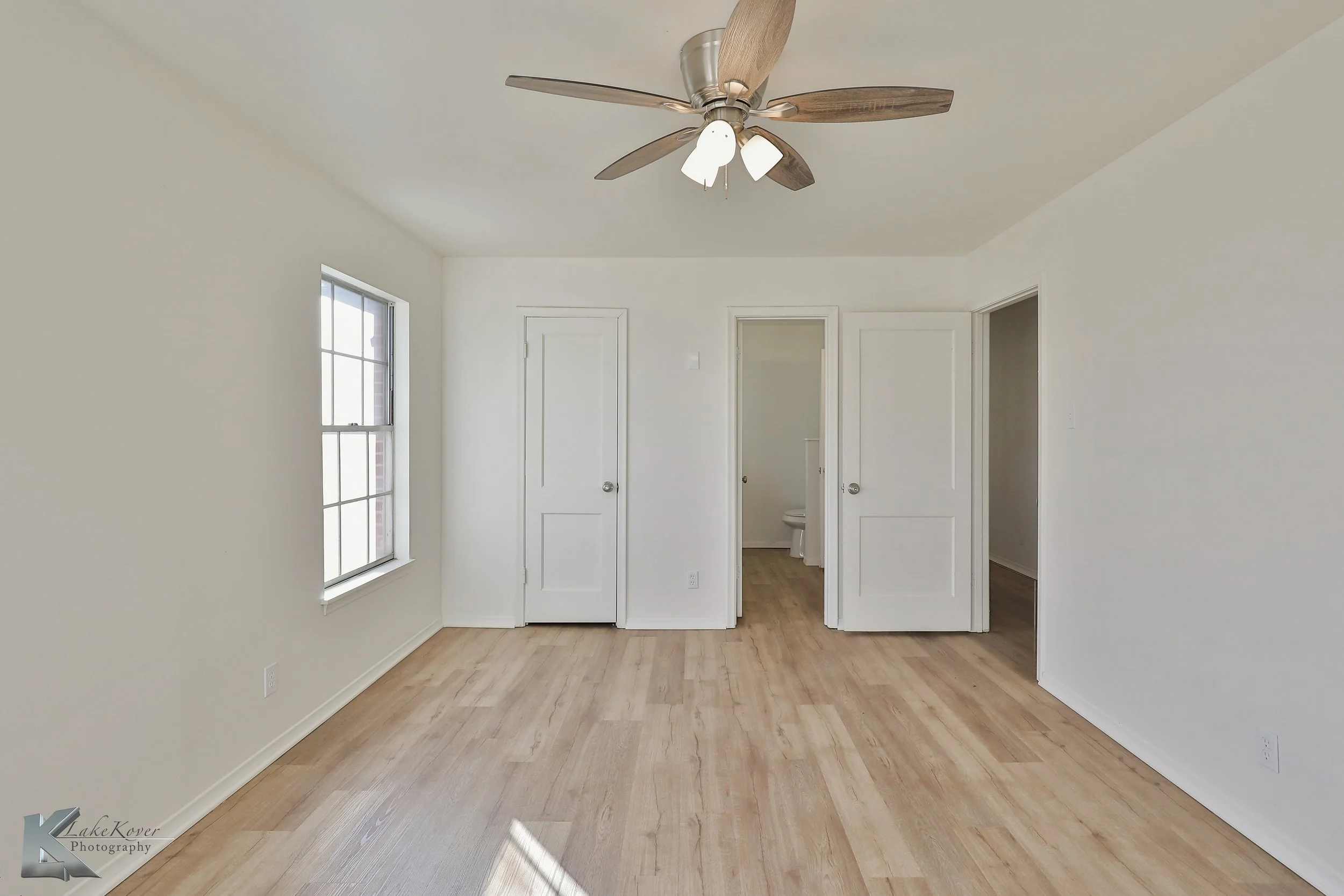 Empty white room with a ceiling fan, two windows, and three doors, one of which leads to a bathroom.