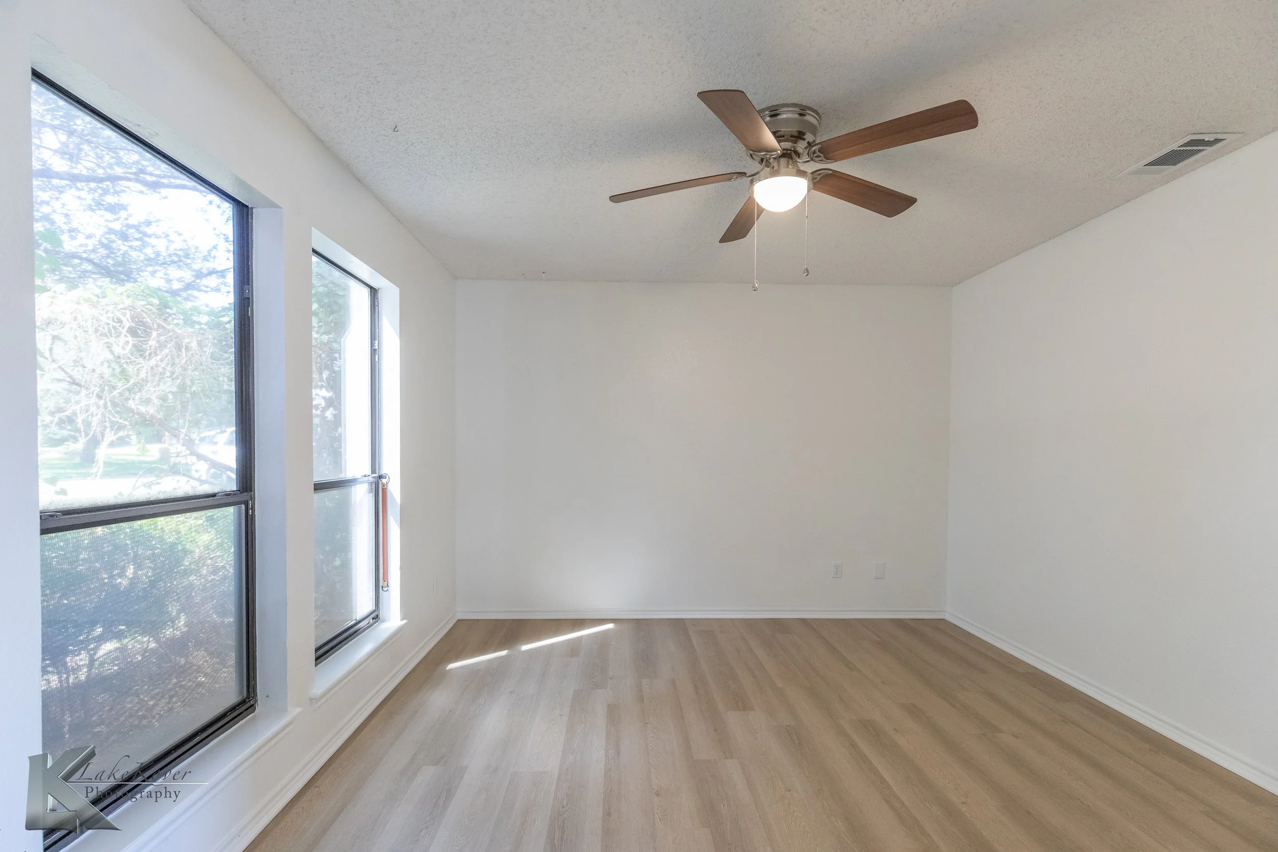 Empty living room with large windows, white walls, wooden floor, ceiling fan, and a light fixture.