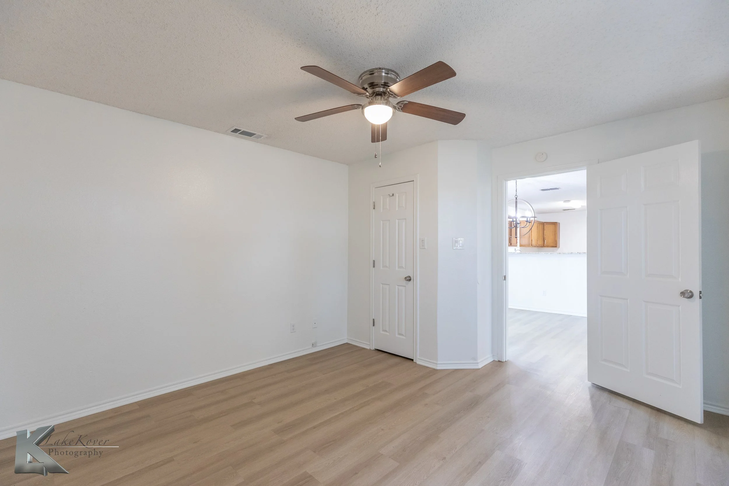 Empty room with white walls, light wood flooring, a ceiling fan with wooden blades, and a door leading to a kitchen area with cabinets and a dining table.