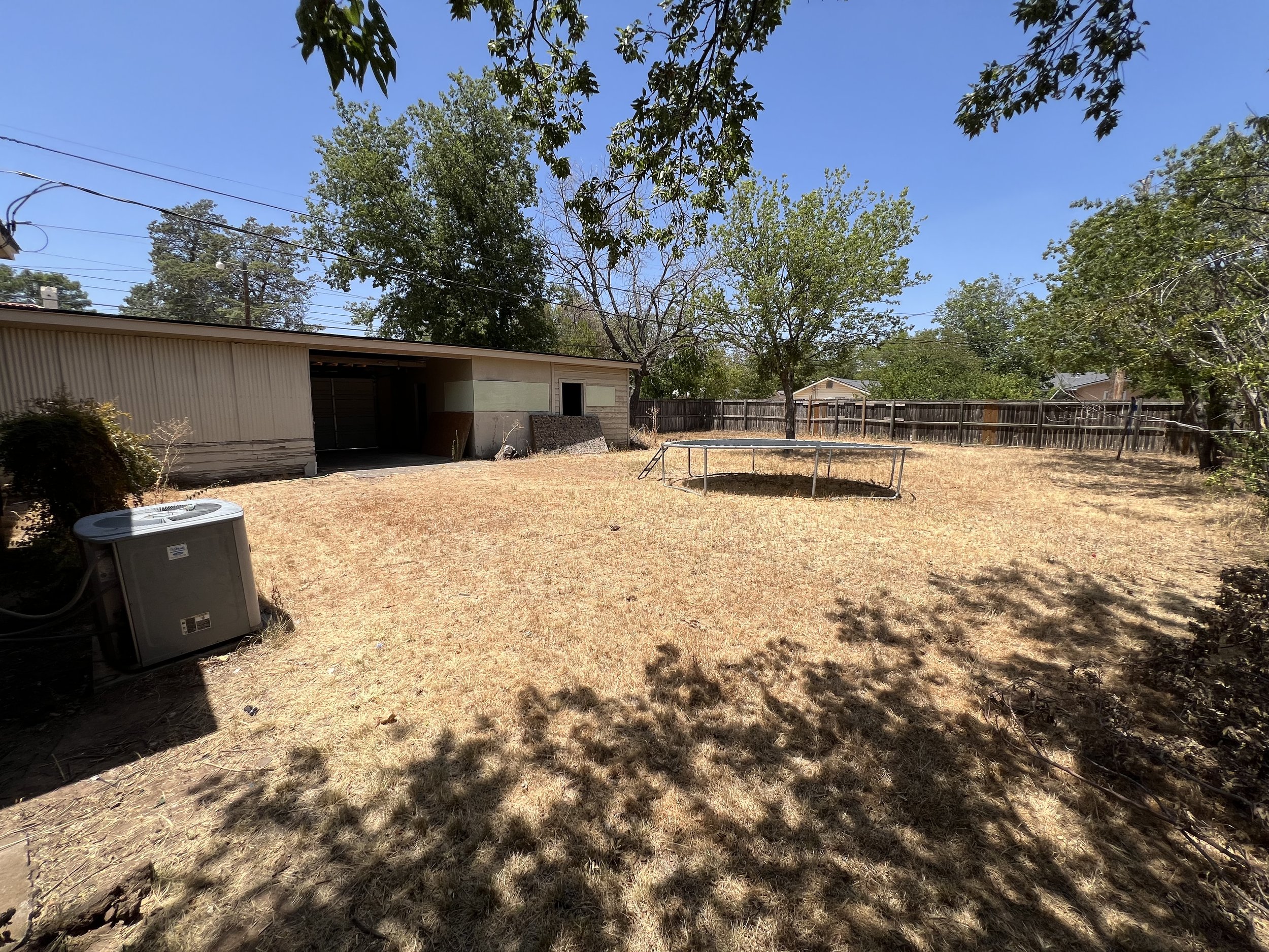 A backyard with dry grass, several trees, a metal shed, a trampoline, and a wooden fence under a clear blue sky.
