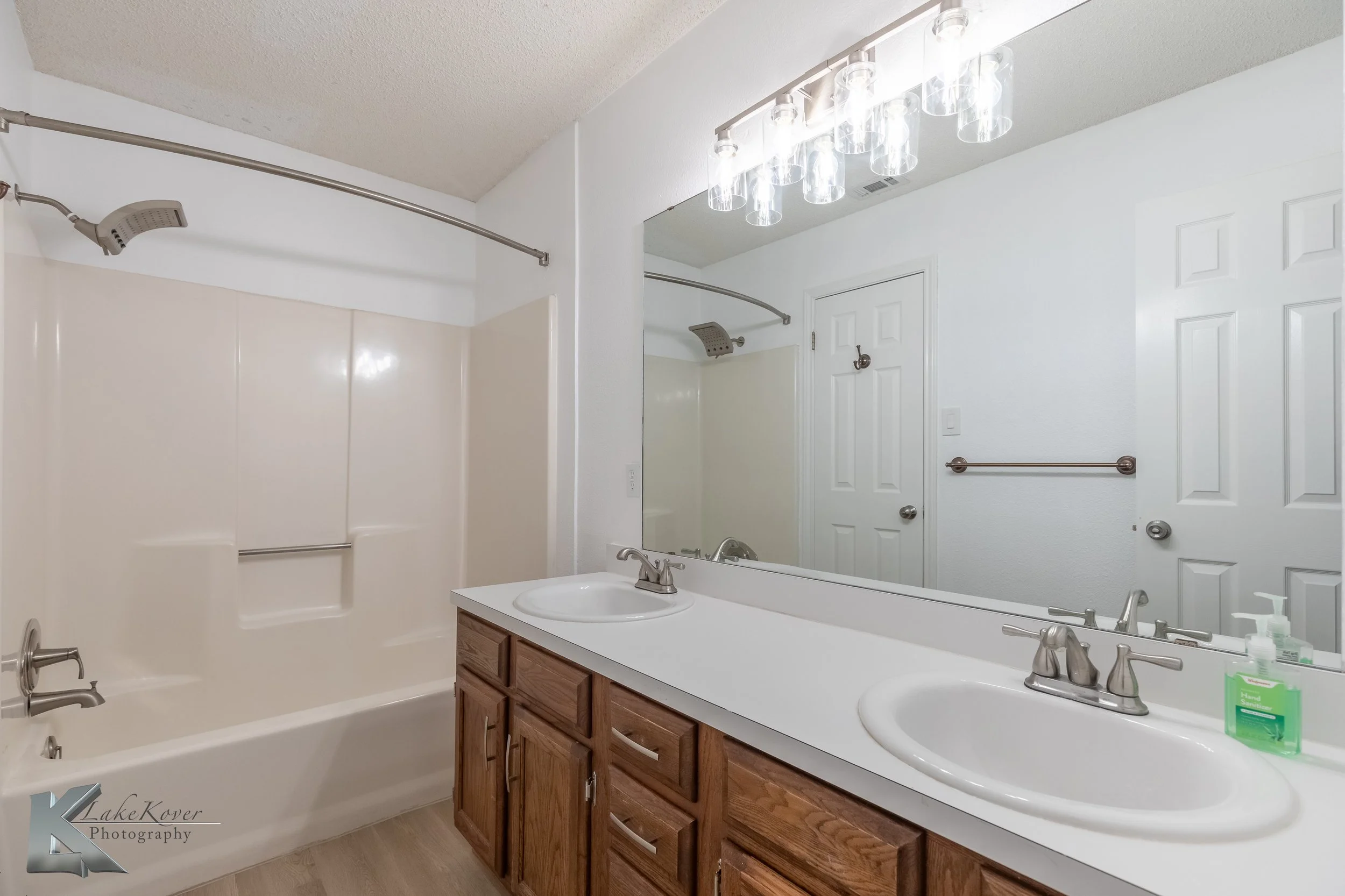 Bathroom with double sink vanity, large mirror, light fixture, white bathtub with shower, and white walls.