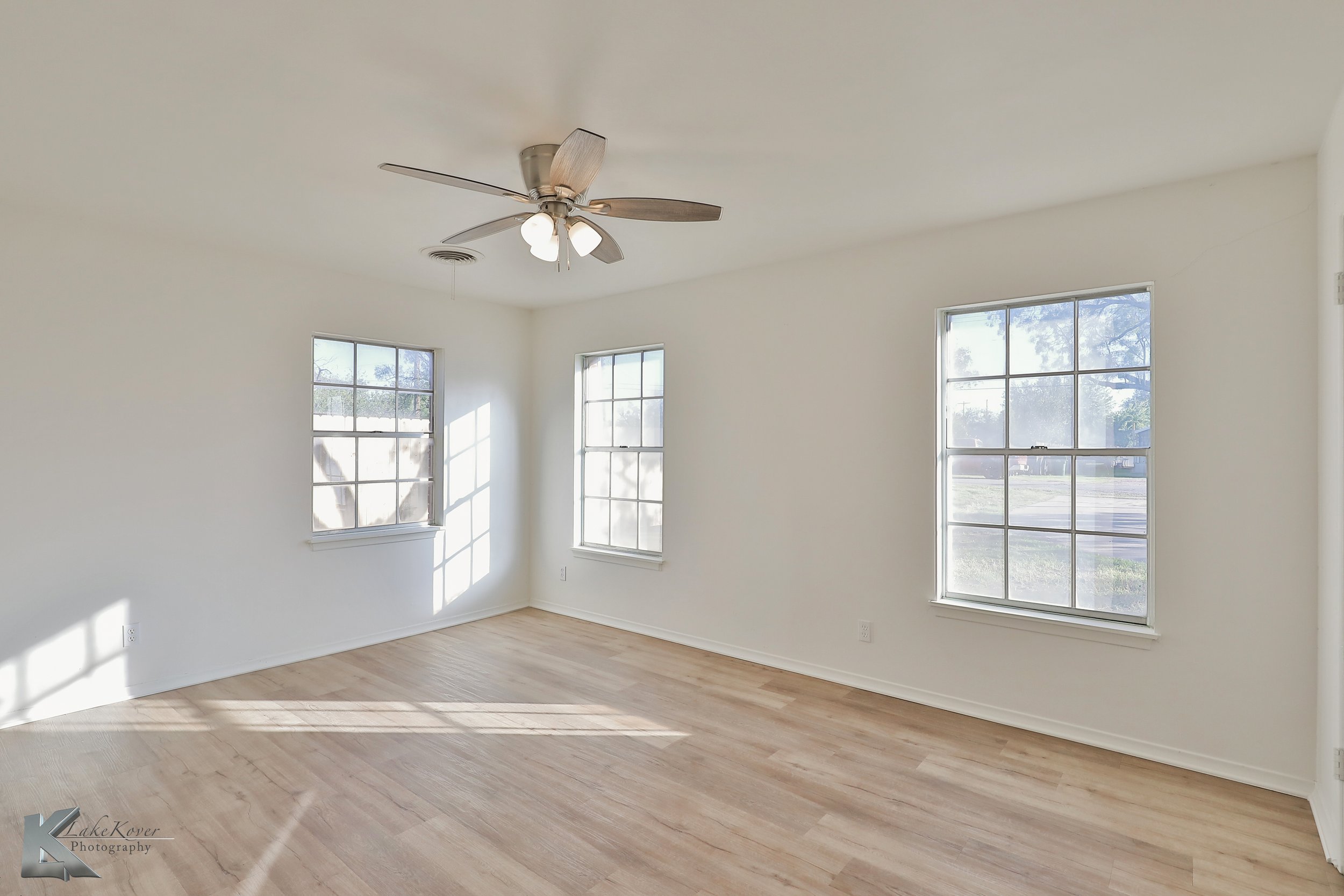 Empty room with white walls, three large windows letting in sunlight, light wood flooring, and a ceiling fan with lights.