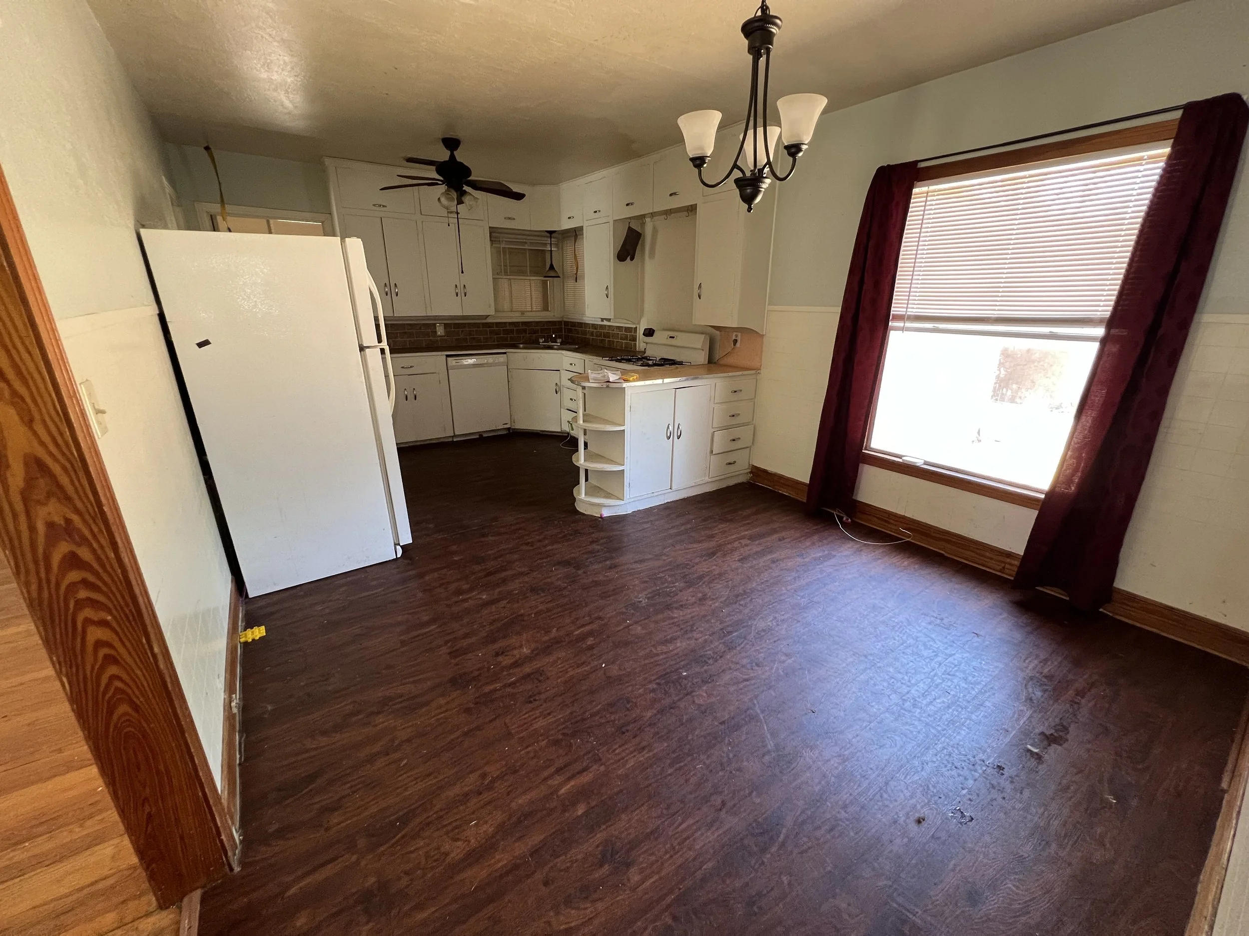 Empty kitchen and dining area with dark wood floors, white cabinets, a window with burgundy curtains, and ceiling light fixtures.