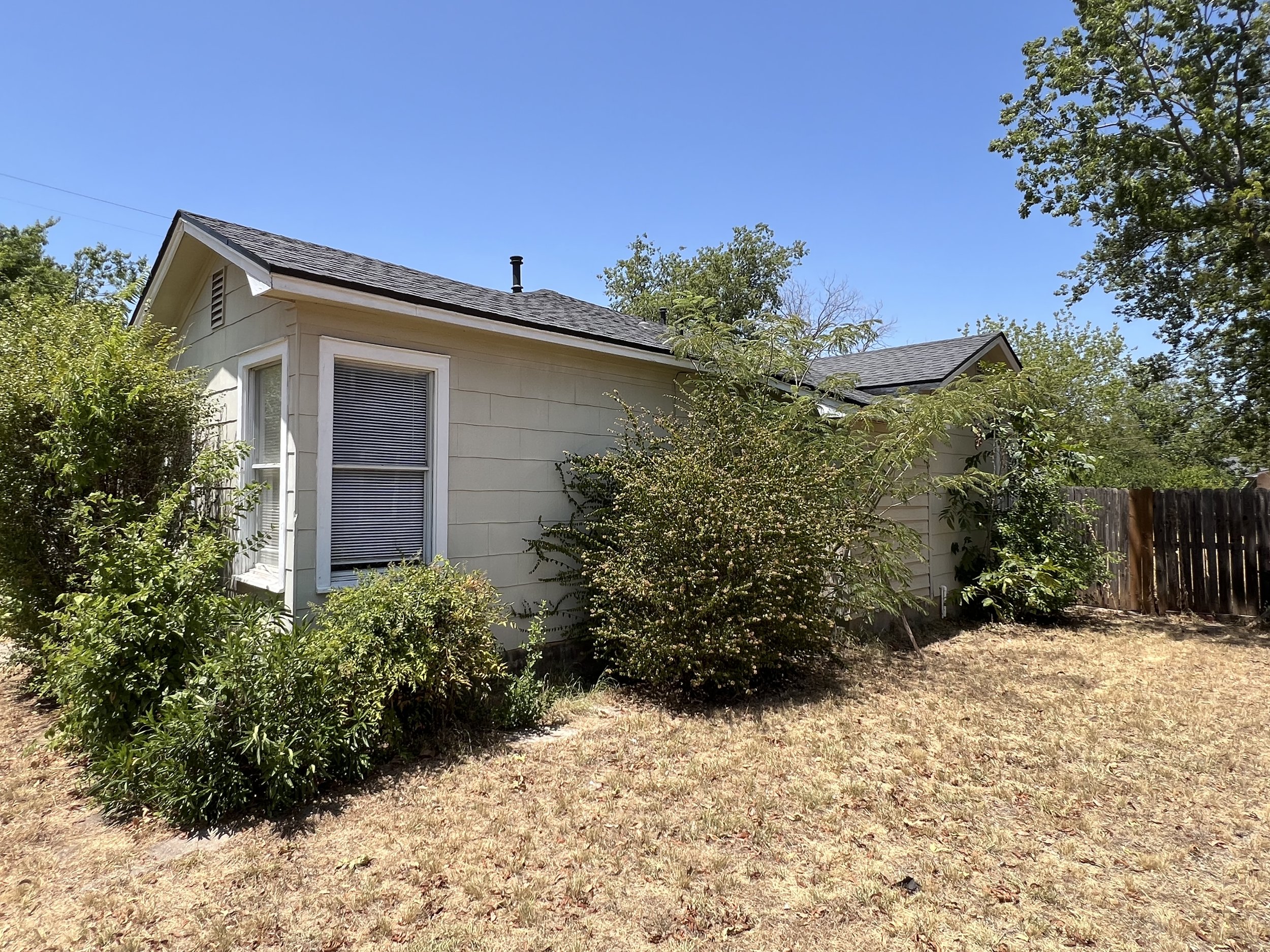 Backyard of a house with cream siding, a window with blinds, bushes along the wall, trees, and a wooden fence under a clear blue sky.