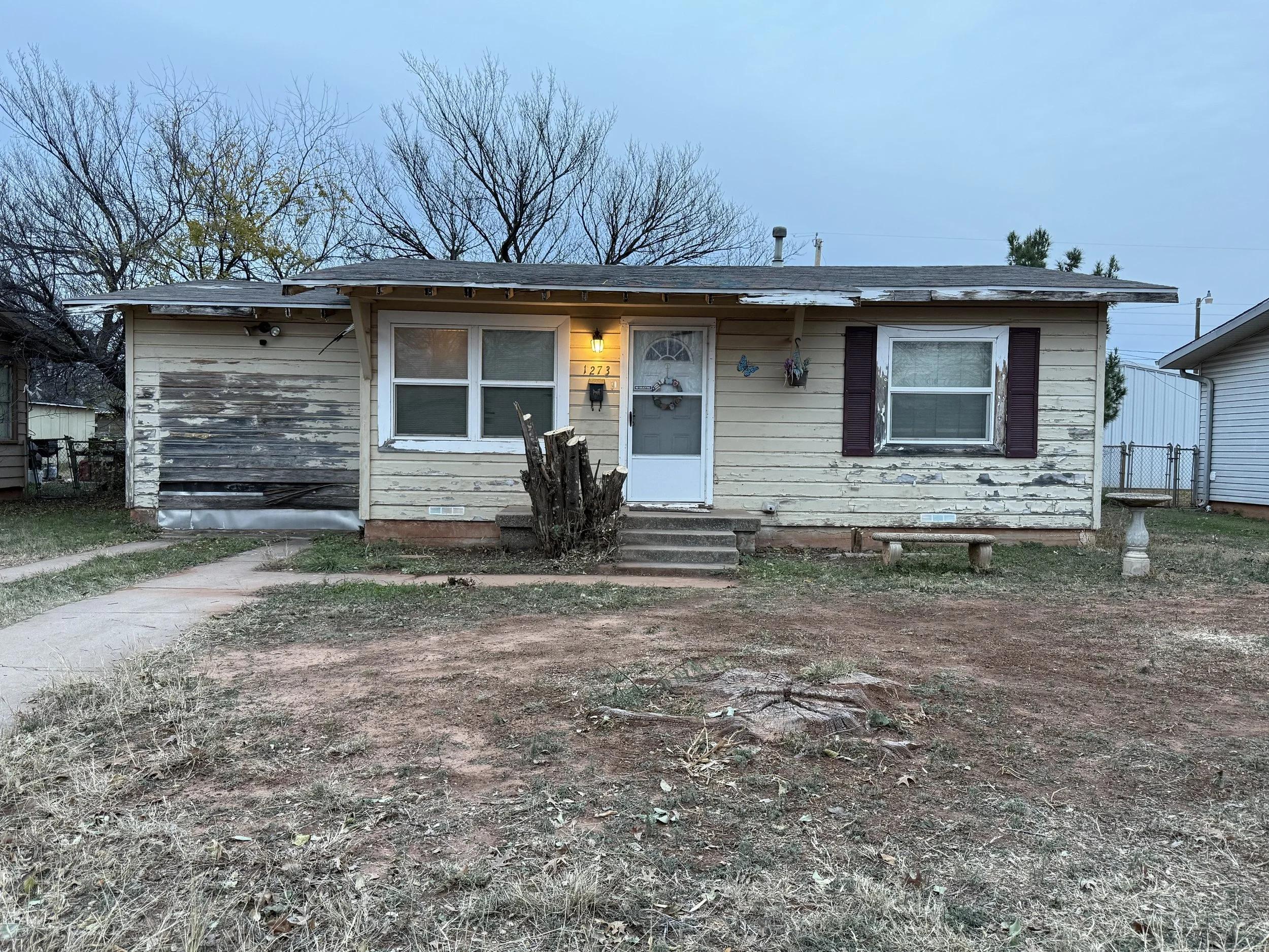 Front view of an old, weathered house with peeling paint, a small porch with steps, and a leafless tree in front. The yard is mostly dirt with patches of grass.
