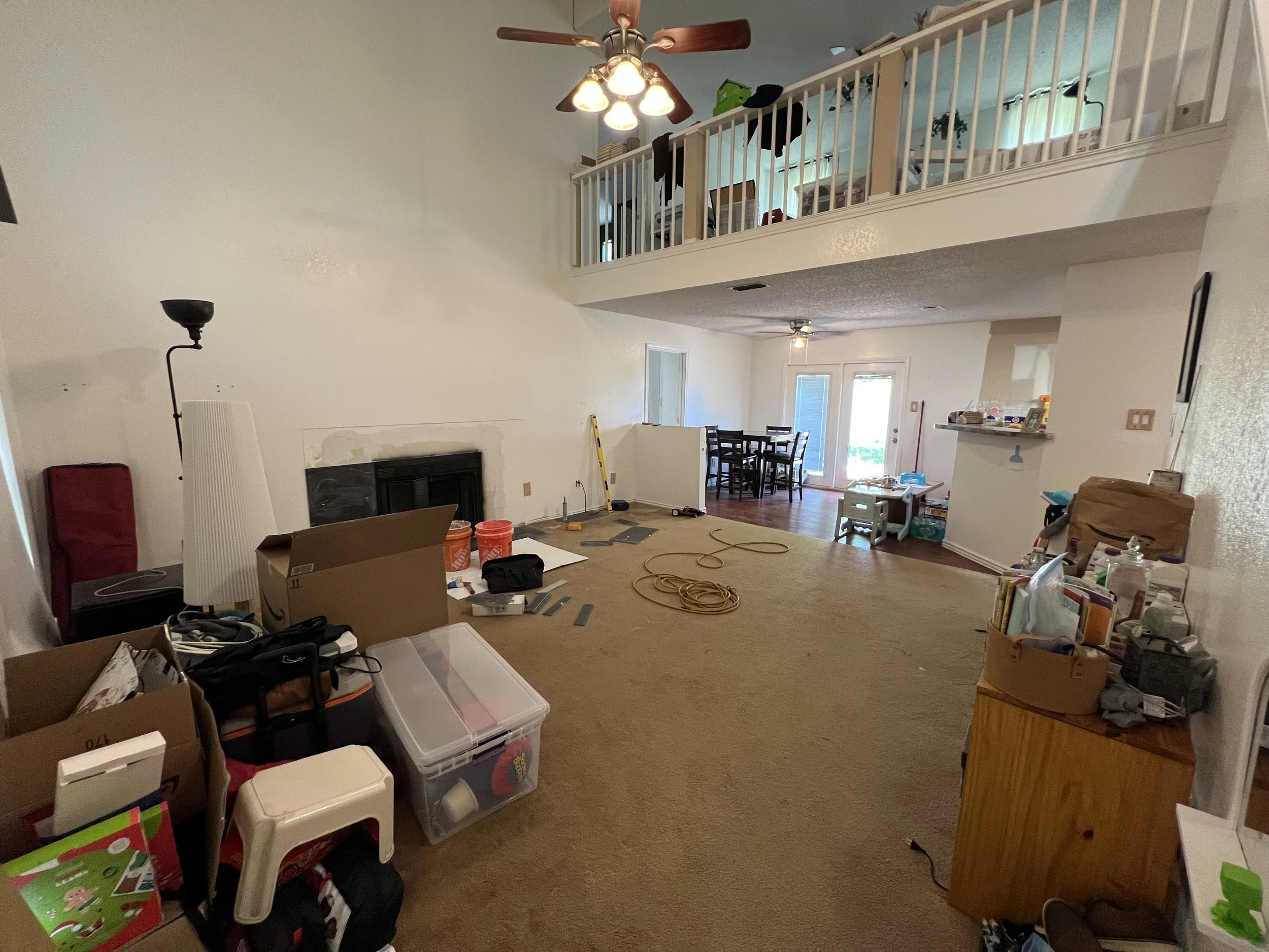 Living room with clutter and unpacked boxes, facing an open floor plan with a dining area and sliding door to outside, with a two-story ceiling and a ceiling fan.