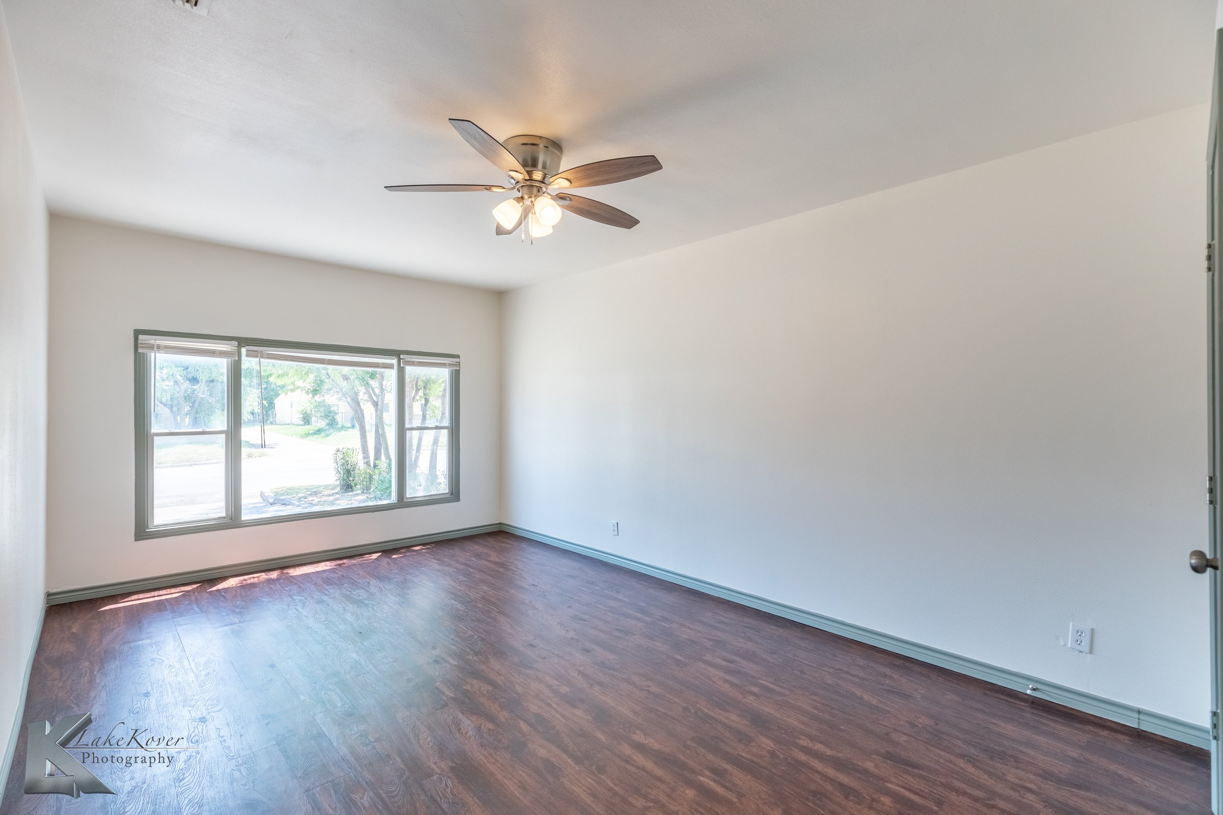 Empty living room with large window, ceiling fan with lights, hardwood floor, white walls, and a door on the right.