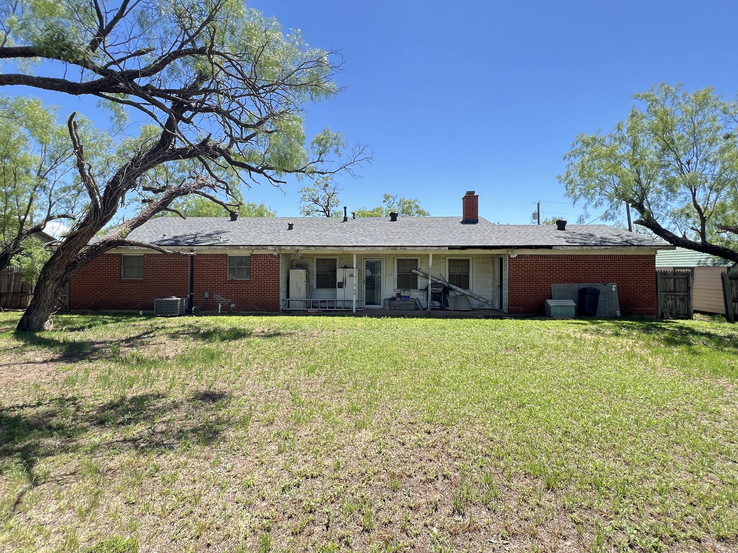 Backyard view of a brick house with trees and a grassy lawn under a blue sky.