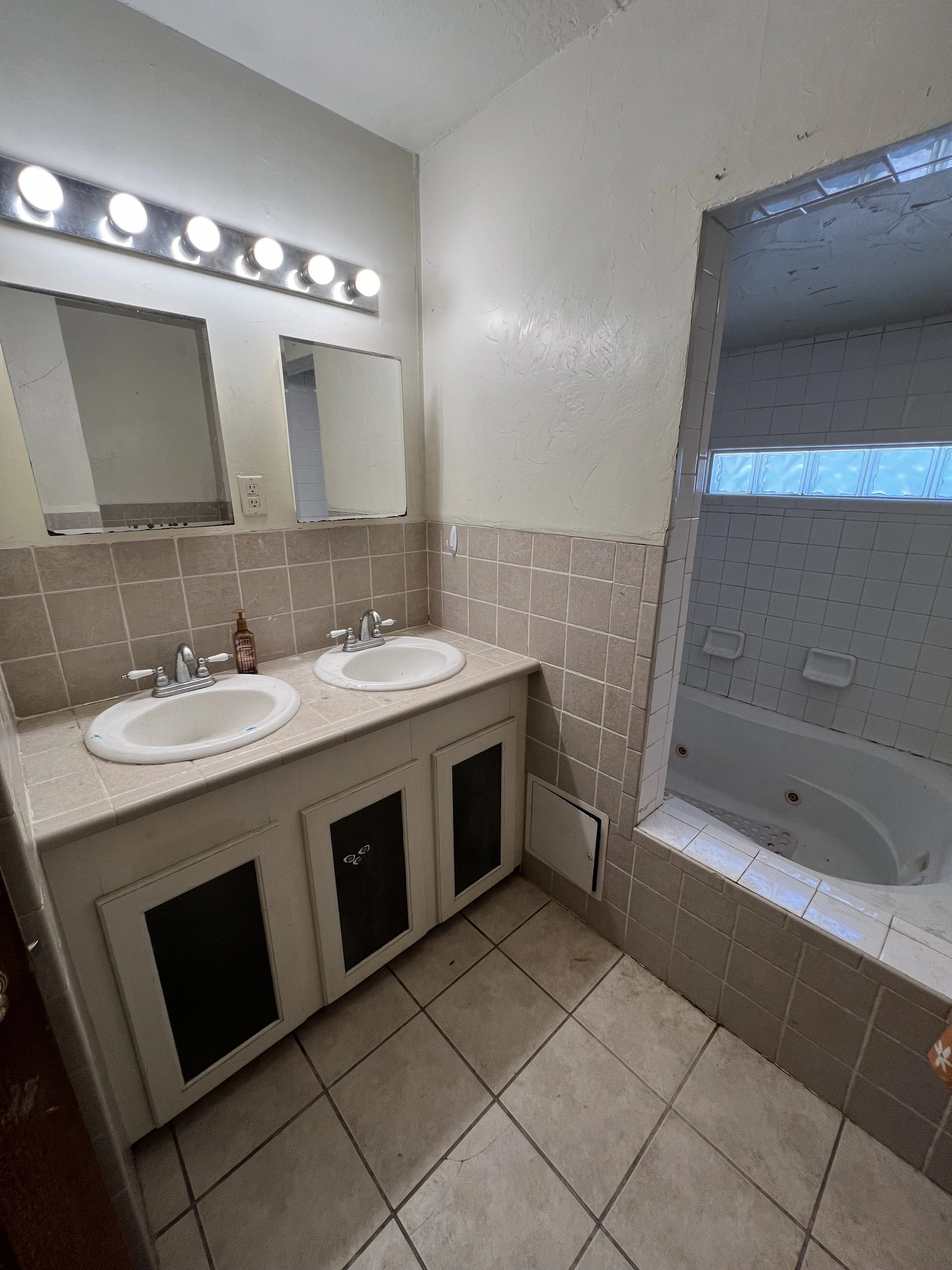 A small bathroom with a double vanity sink, mirror, and a wall of lights above it, next to a shower with a frosted glass block window.