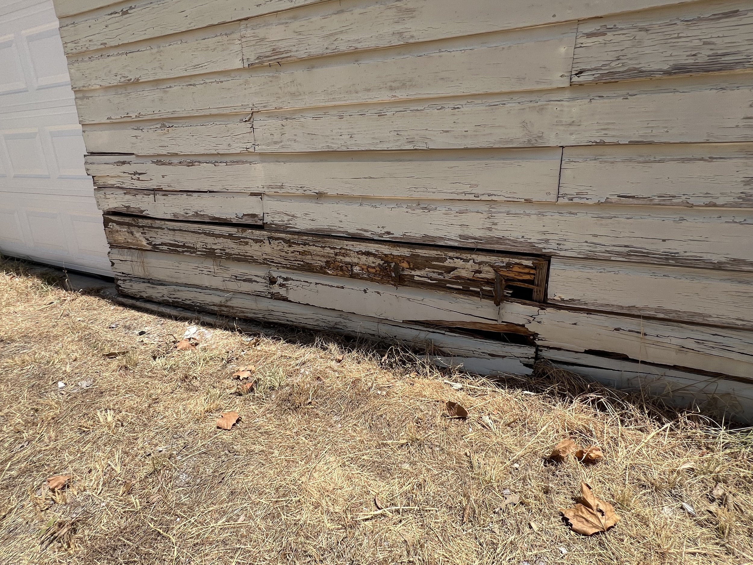 A house with peeling white paint on its wooden siding, some boards are cracked and a section near the bottom is rotting and damaged.