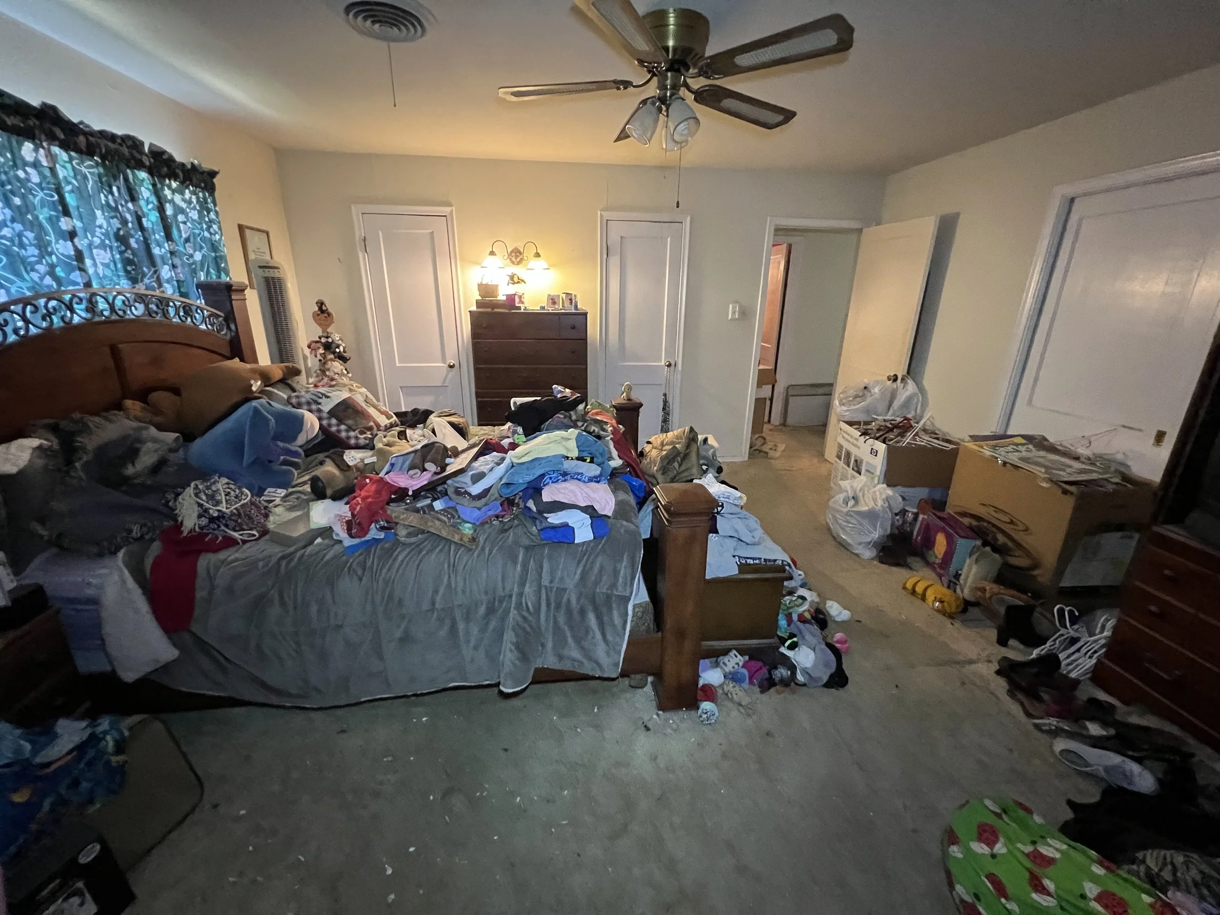 Unkempt bedroom with a large bed covered in clothes, pillows, and miscellaneous items; cluttered floor with shoes and toys; dresser with lamps and personal items; open closet and doorways; ceiling fan; window with floral curtains.