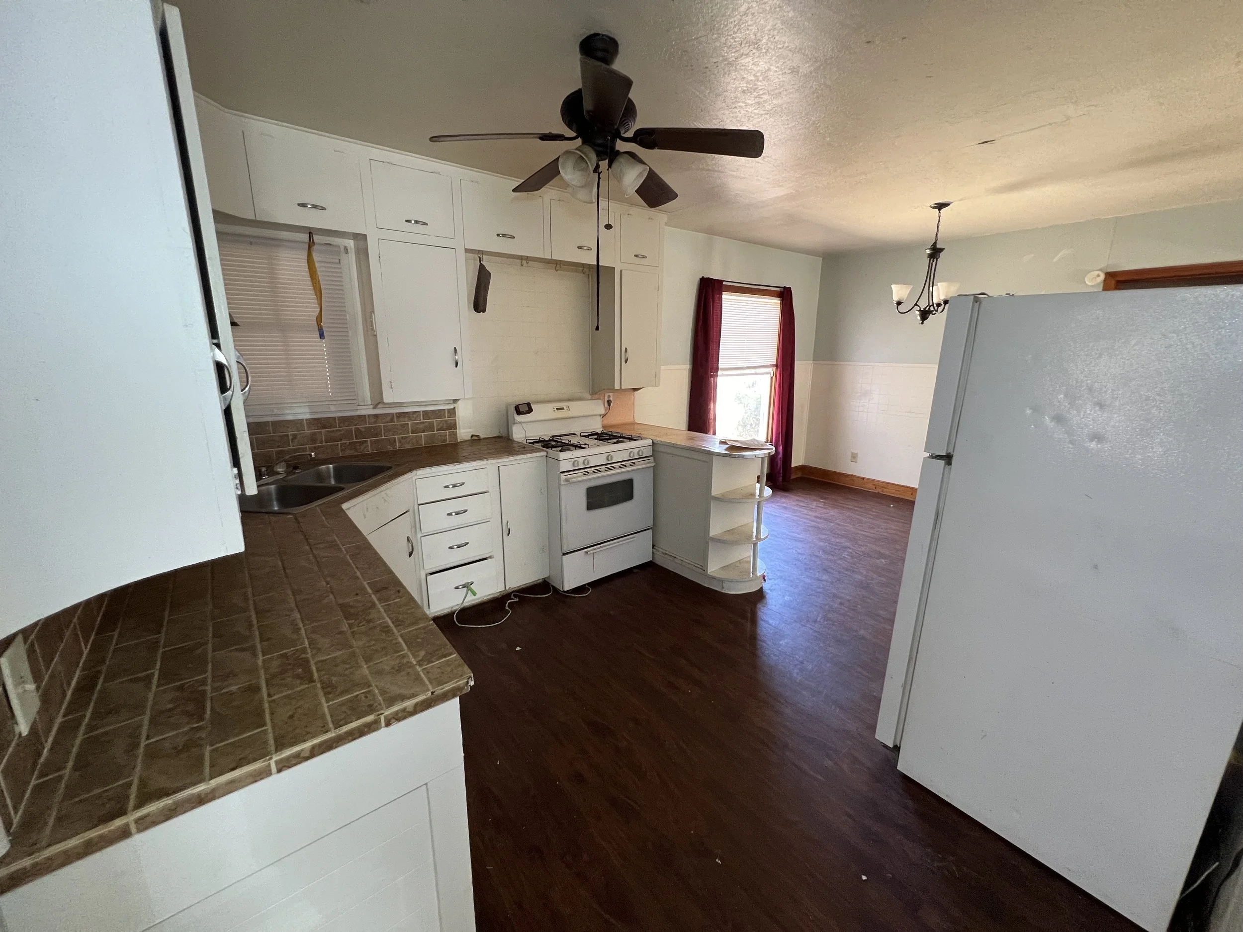 Empty kitchen with white cabinets, brown tiled countertops, a white stove, a white refrigerator, a double sink, dark wood flooring, a ceiling fan, and a window with red curtains.