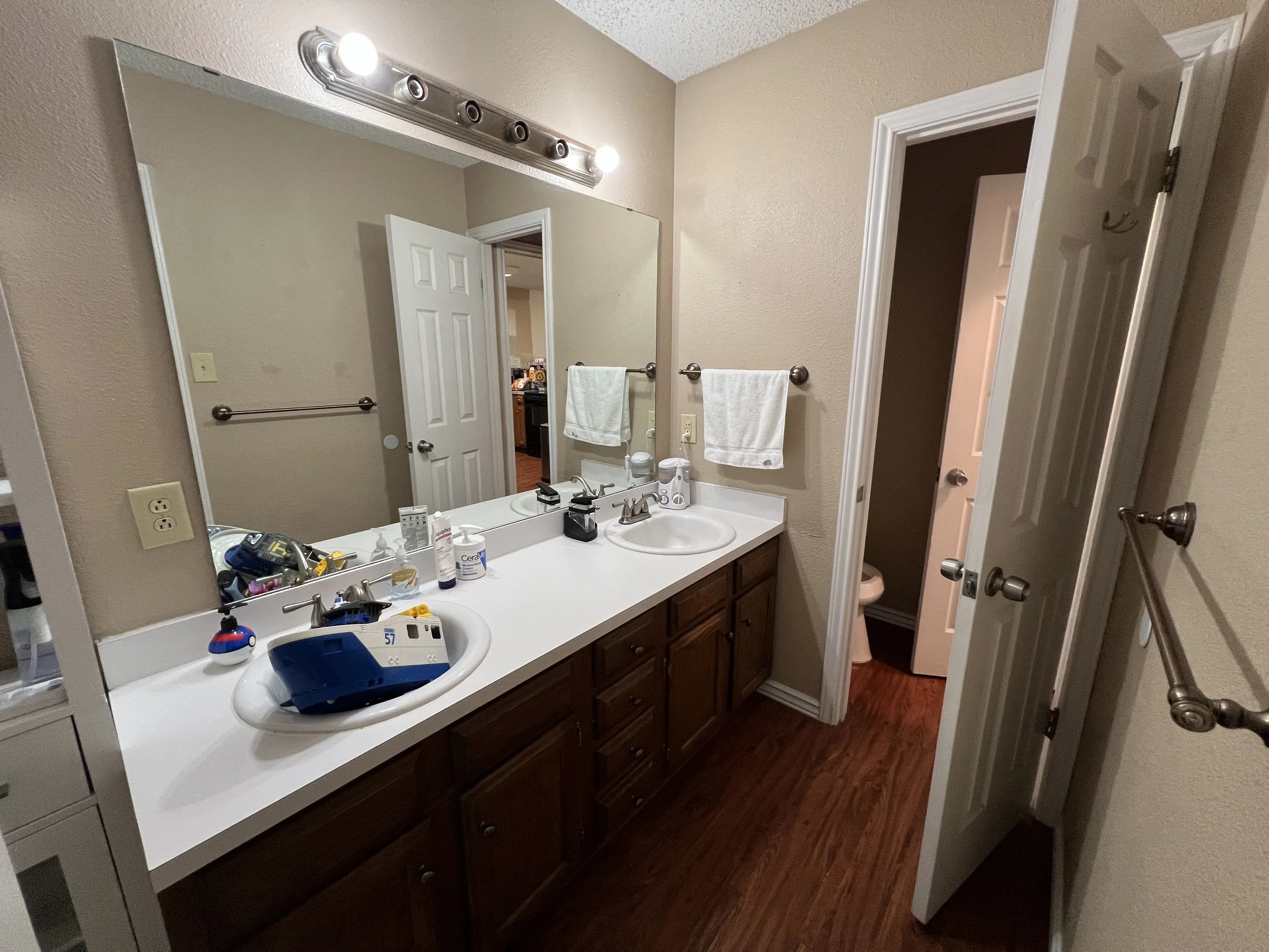 Bathroom with double vanity sink, large mirror, towel racks, and a small closet or toilet room behind a door. The countertop has various toiletries and a blue device. The floor is wooden, and the walls are beige.