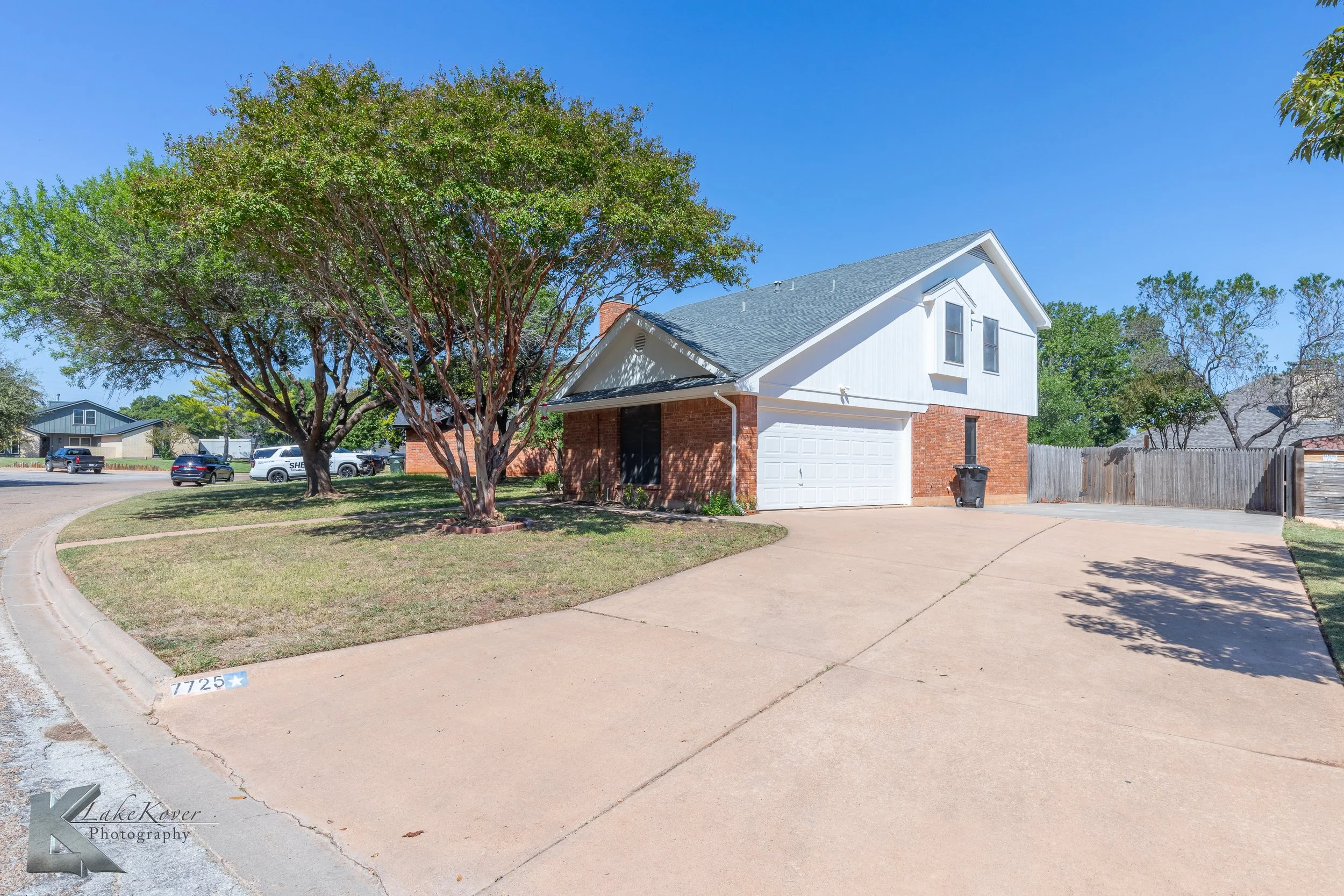 A two-story house with red brick on the lower level and white siding on the upper level, with a gray roof, garage, large tree, and a curved driveway in a suburban neighborhood.