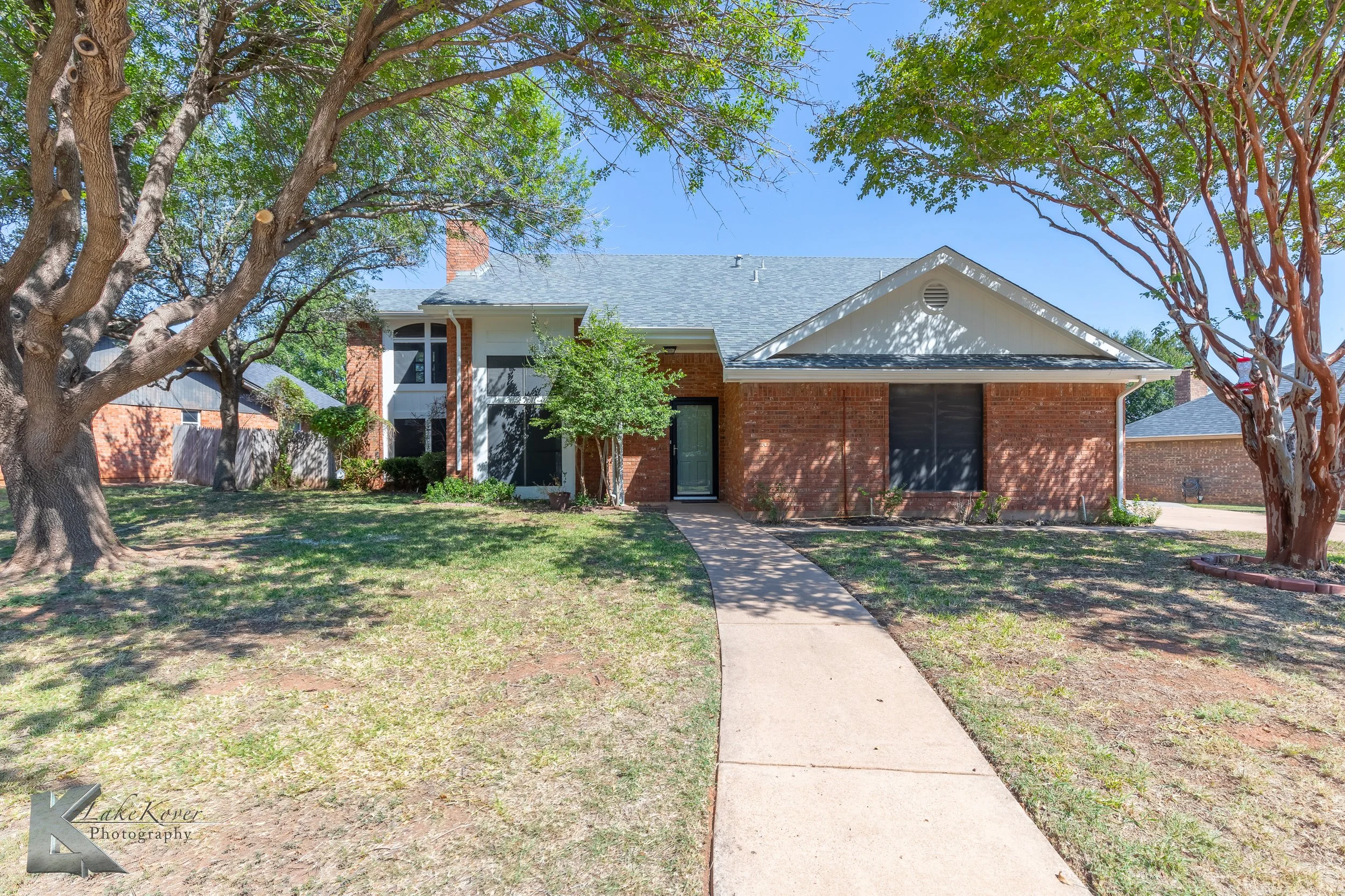 Front view of a two-story house with red brick and white siding, black front door, large windows, a concrete walkway, surrounded by trees and a lawn, sunny day.