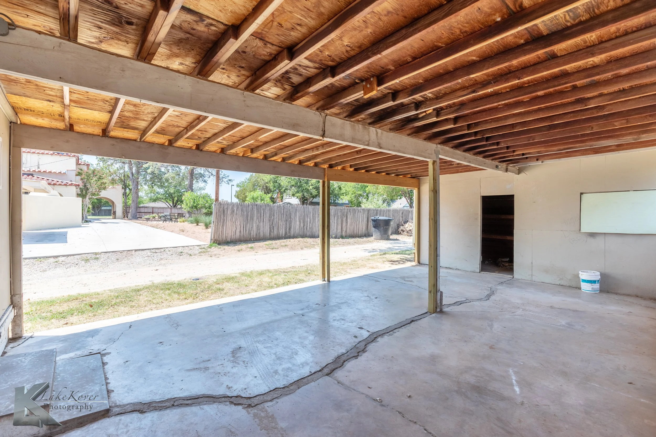 Under-construction backyard patio with concrete floor, open view to outdoor yard, wooden ceiling beams, and a small storage area