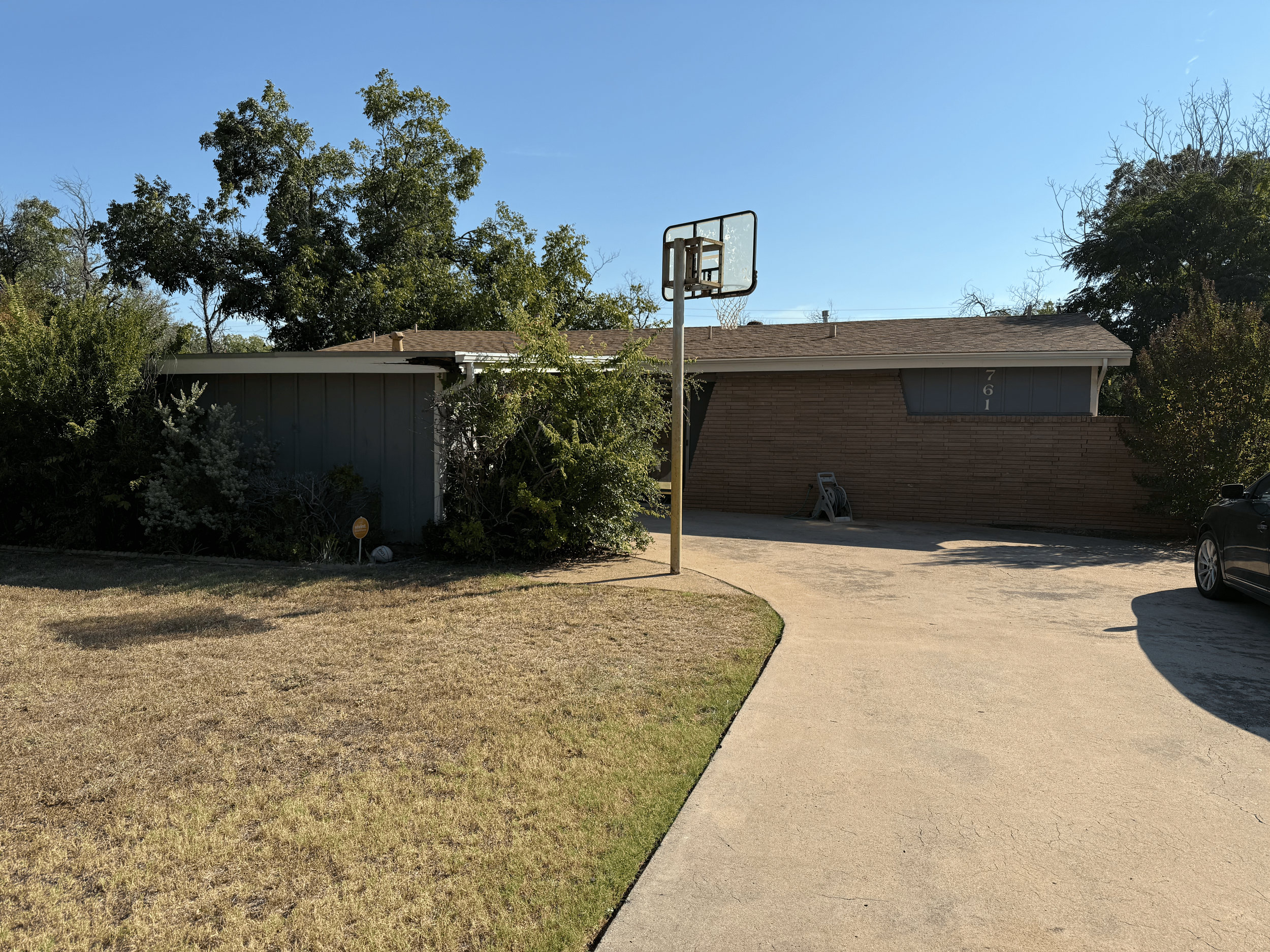 A house with a tennis hoop in the driveway, a black car parked on the right, and trees in the yard, under a bright blue sky.
