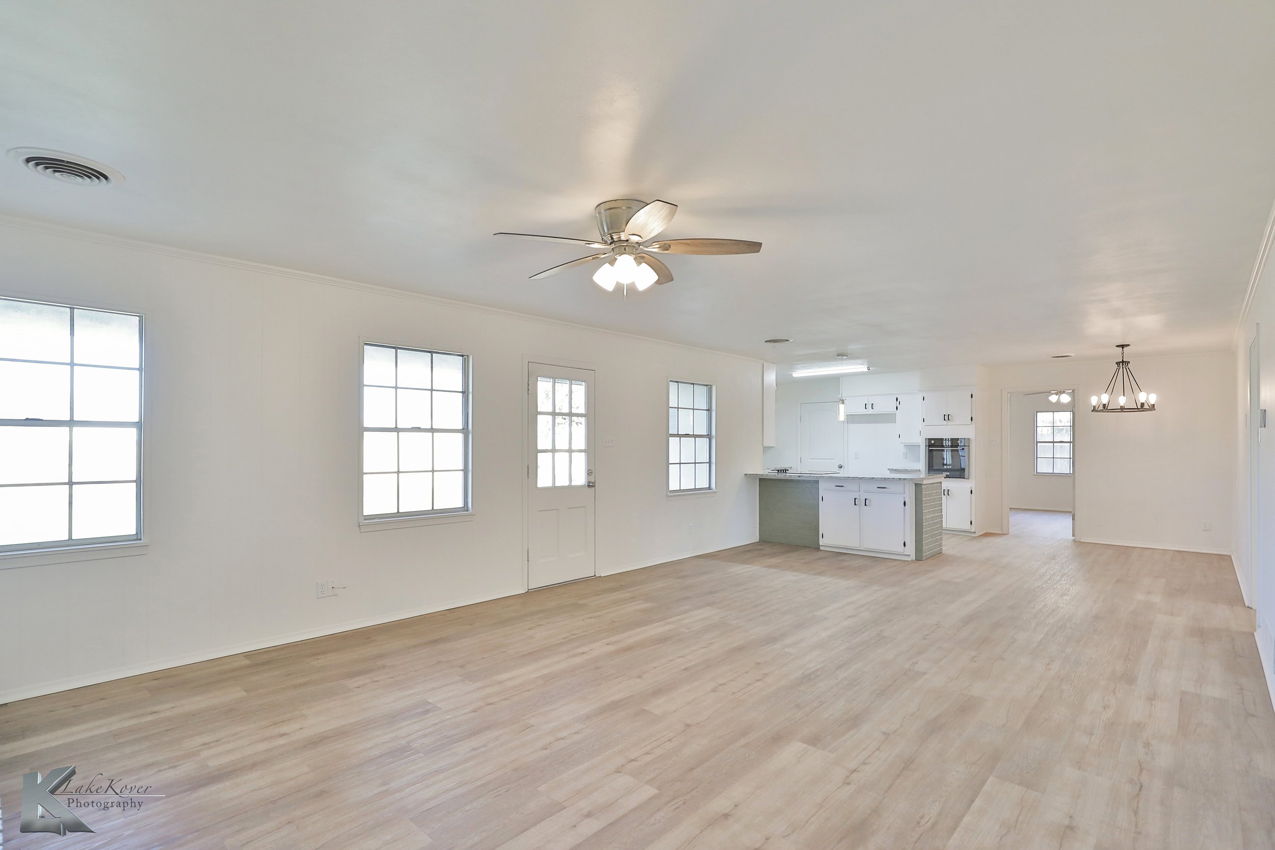 Empty living room and kitchen with white walls, hardwood flooring, ceiling fan, multiple windows, and a chandelier.