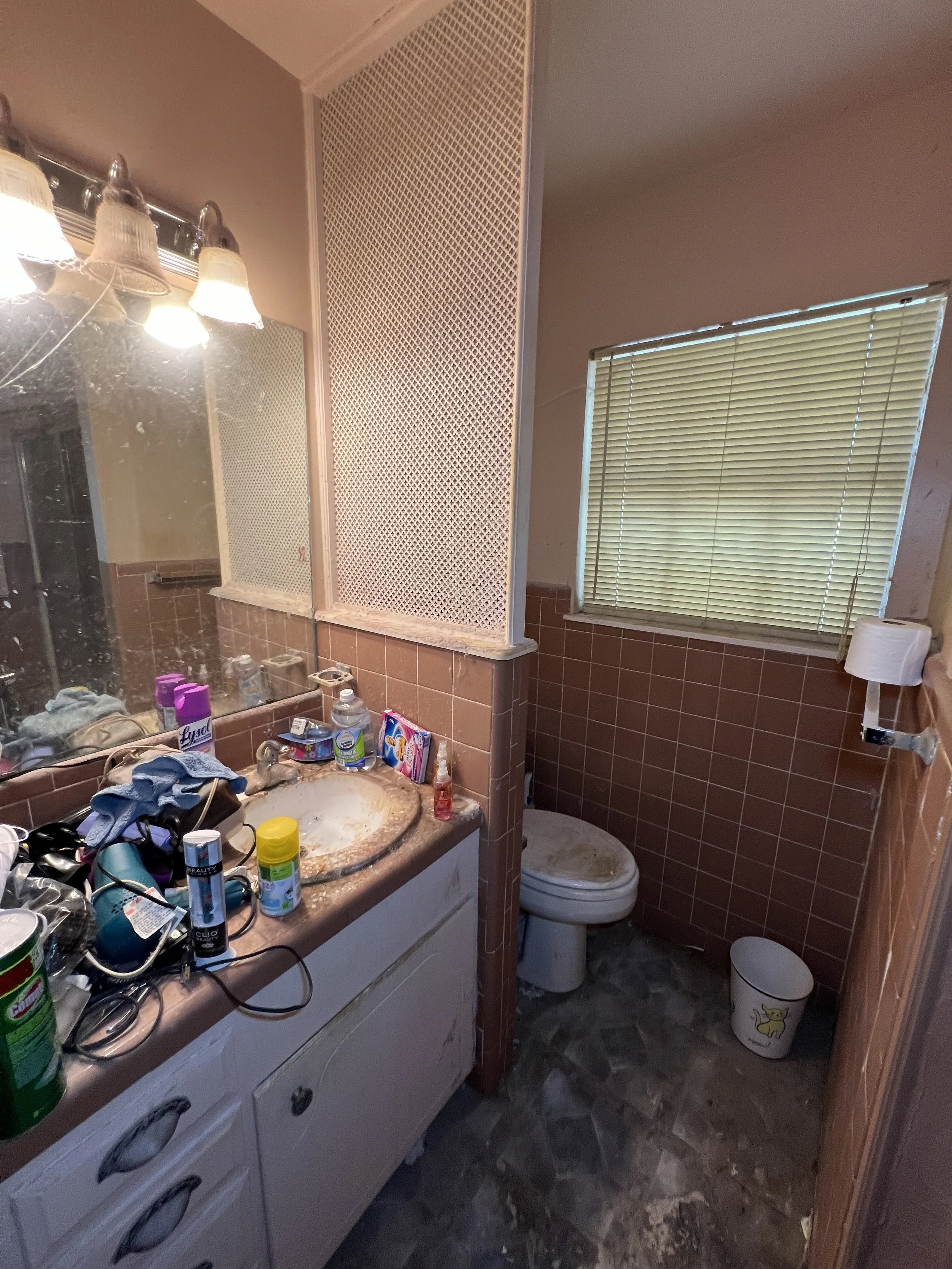 A cluttered bathroom with a dirty sink, messy counter, closed window with blinds, and a toilet with a dirty lid. The mirror above the sink is dirty, and the room has a dim, unclean appearance.