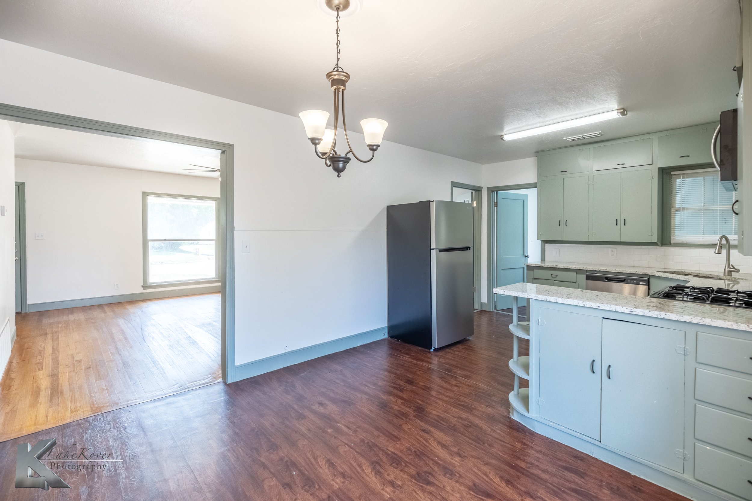 Kitchen with light blue cabinets, white countertops, stainless steel appliances, and a ceiling chandelier. Adjacent room has a large window and hardwood floors.