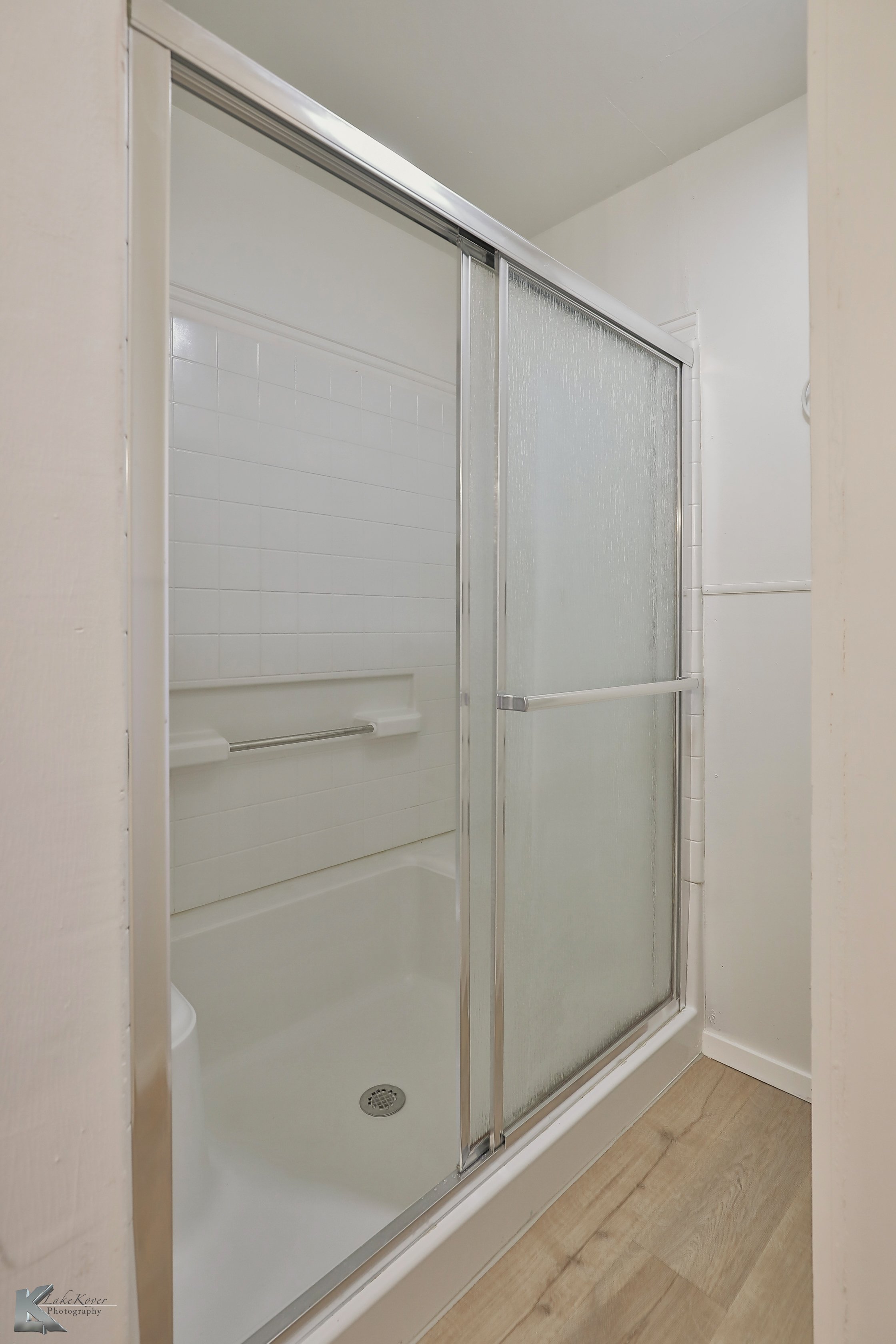 Bathroom shower with a sliding glass door, white tiled wall, and a grab bar inside.