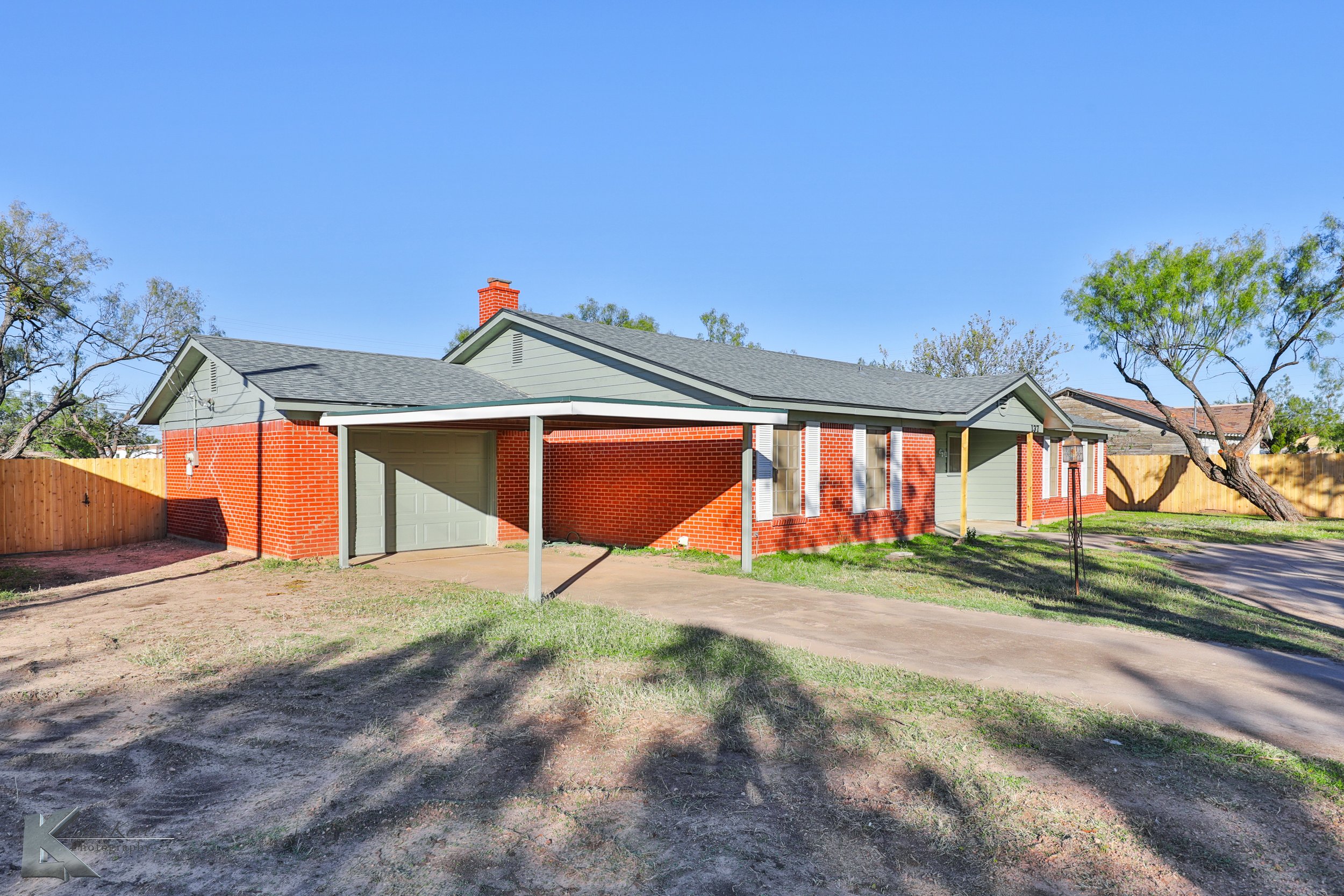 Single-story suburban house with red brick walls, gray roof, and attached garage, surrounded by a fenced yard and trees under a clear blue sky.