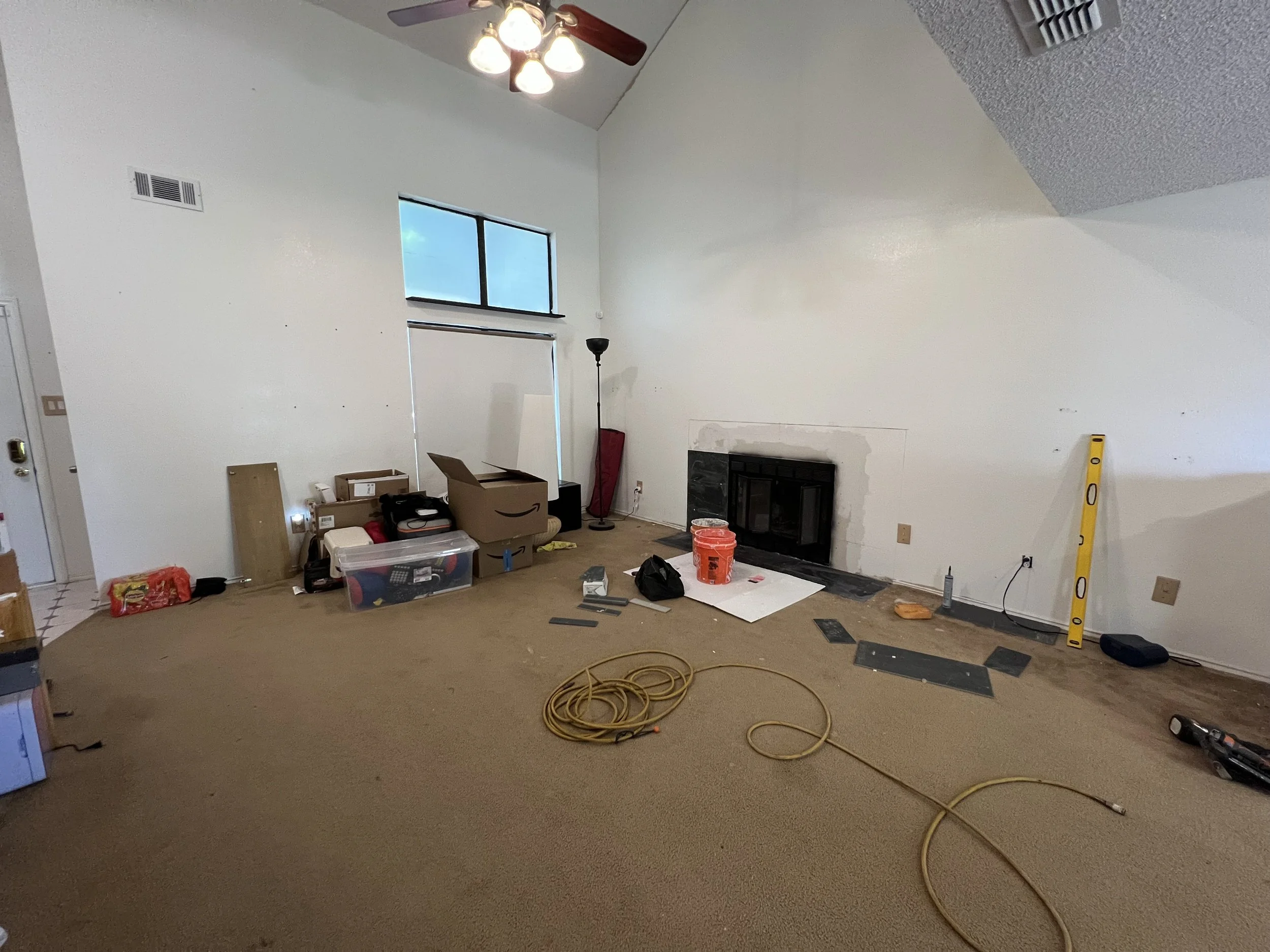 Living room room under renovation with tools, construction materials, and furniture moved aside. Visible fireplace, window, and ceiling fan with lights.