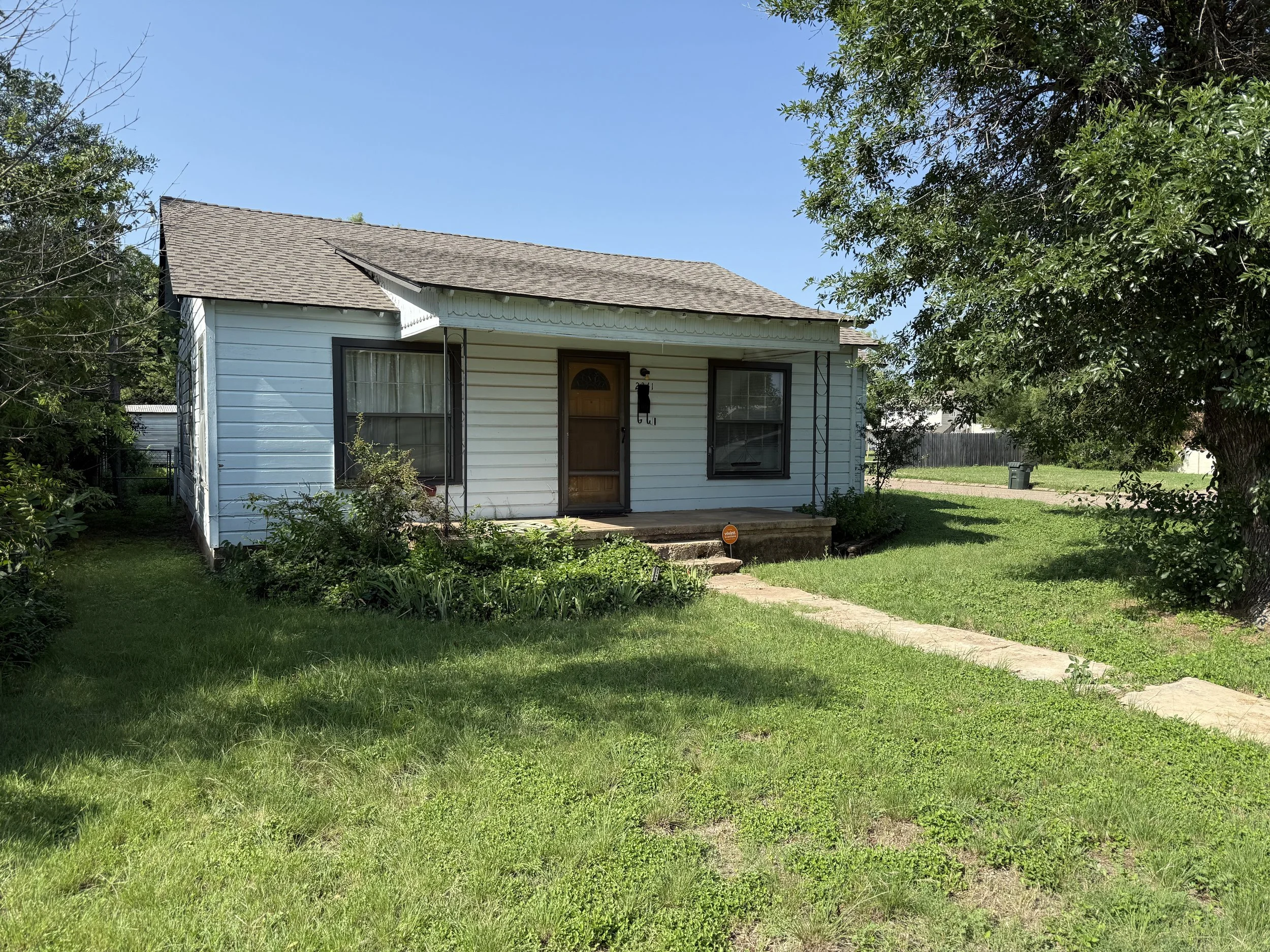 Small white house with black trim, front porch, and steps leading to a green lawn, with trees and clear blue sky in the background.