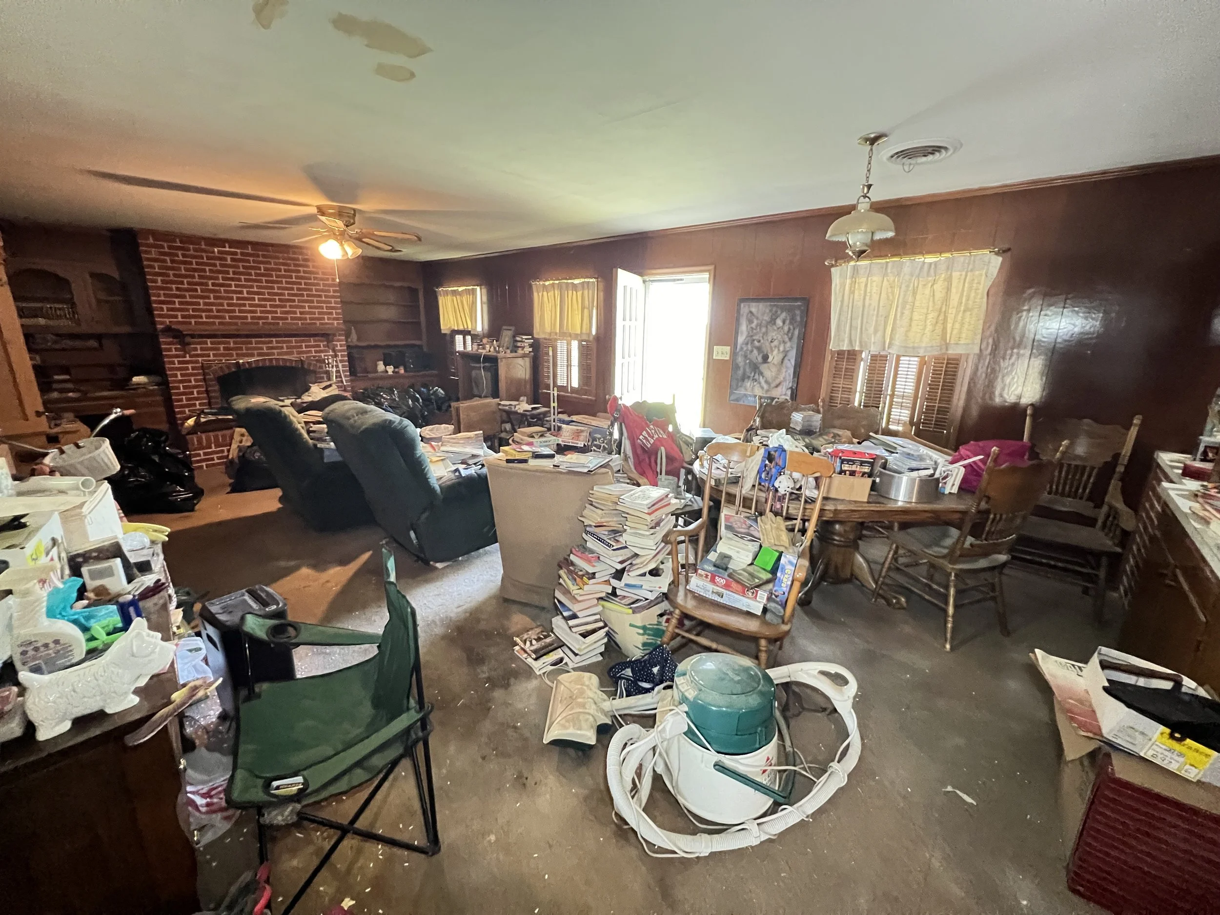 A cluttered living room with piles of books and papers on furniture, a green folding chair, a vacuum cleaner, two black recliners, and a brick fireplace. There are windows with yellow curtains, a painting of a lion, and various items scattered across