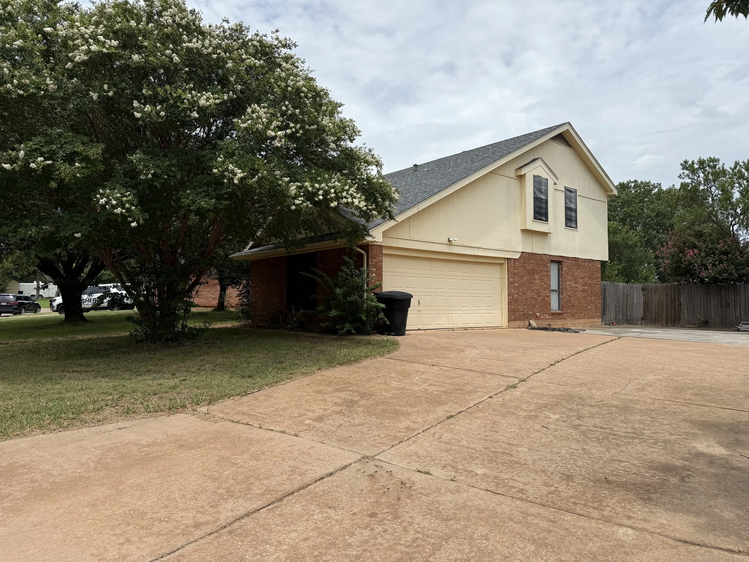 A two-story house with a brick and beige exterior, a closed garage door, and a large leafy tree in the front yard. The driveway is concrete with some cracks, and there is a black trash bin near the garage. Fencing and plants are visible at the back a