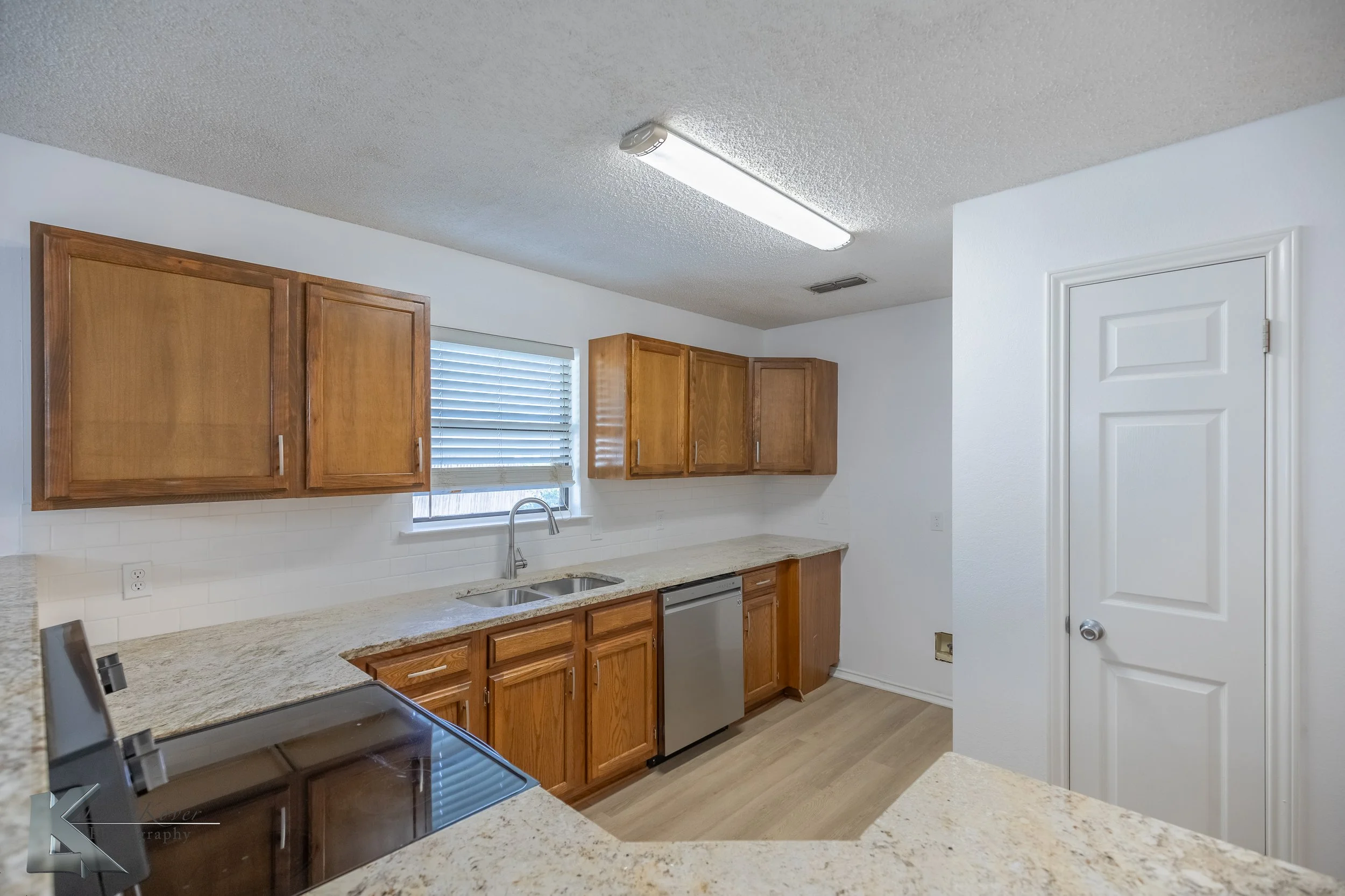 Kitchen with wooden cabinets, granite countertops, stainless steel dishwasher, white walls, and a window with blinds.
