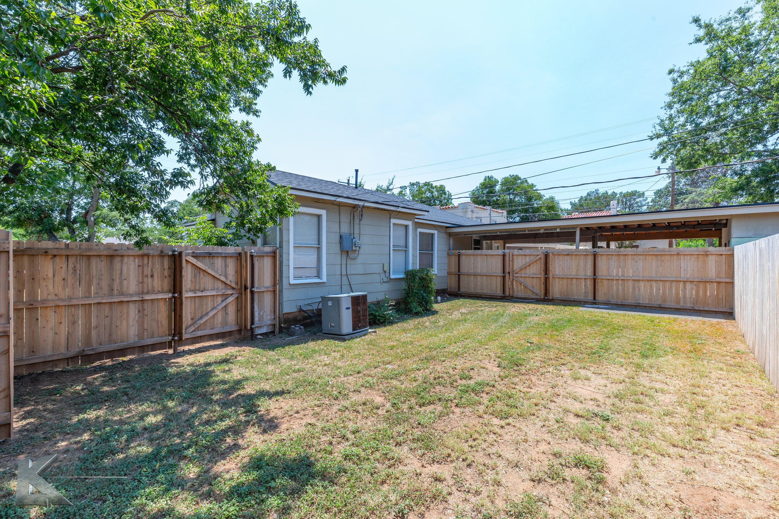 A backyard with short grass, enclosed by a wooden fence, with a small tree and house with windows and air conditioning units visible.
