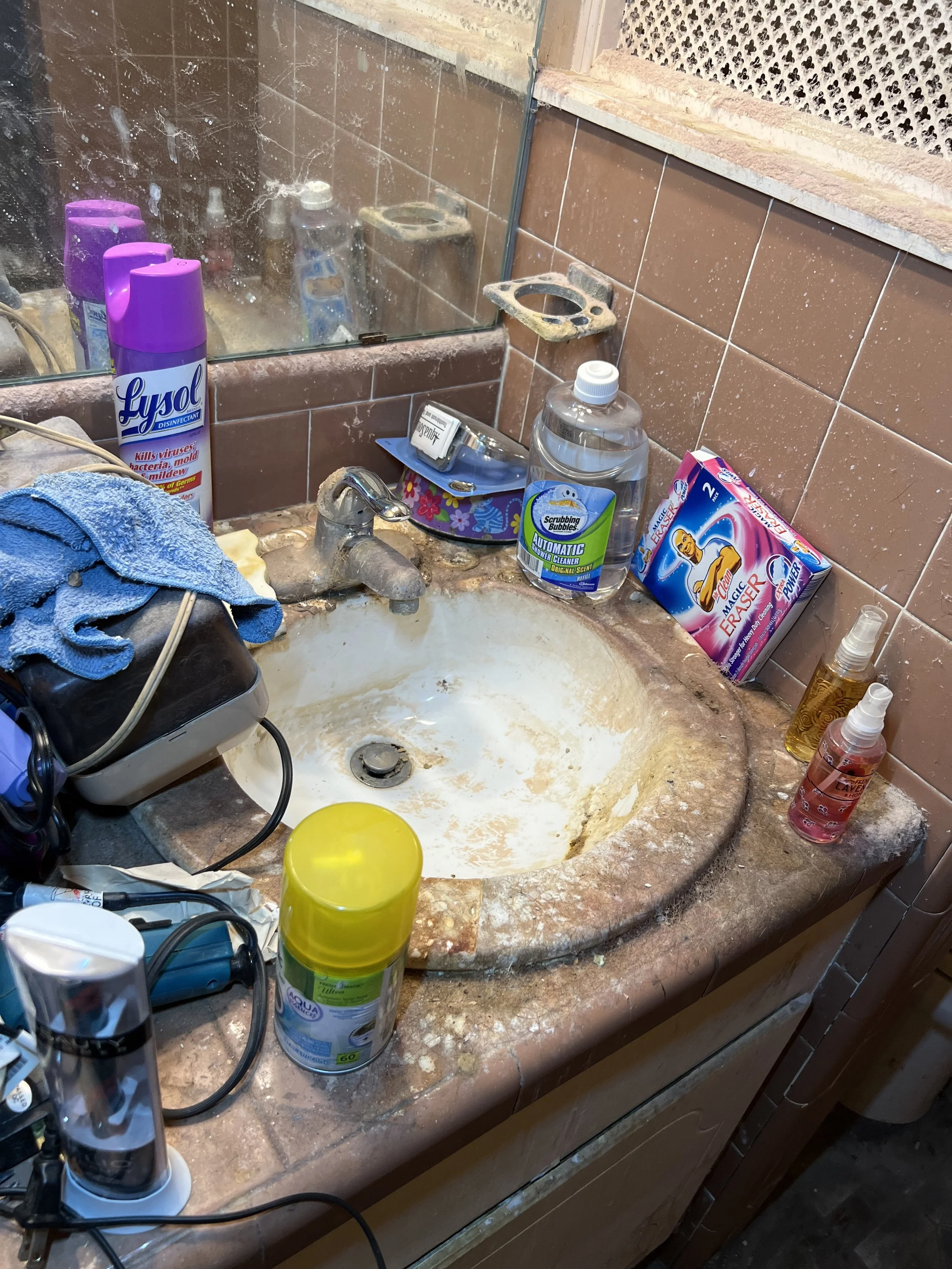 Messy bathroom sink with dirty soap scum, used toiletries, cleaning supplies, and cluttered items around the sink.