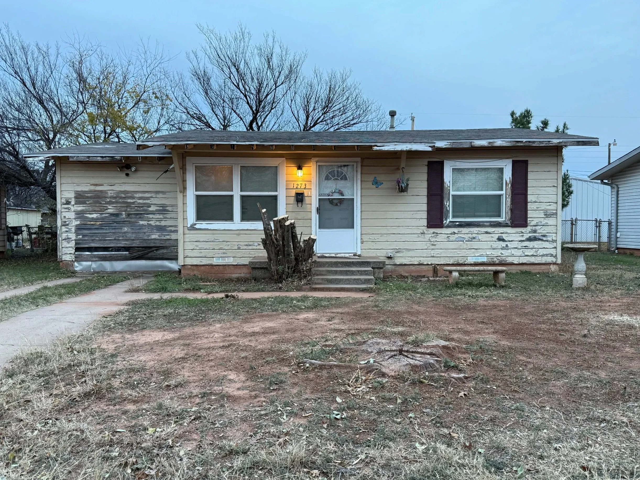 Front of an old, weathered house with peeling paint, a lit porch light, and leafless trees in the background during dusk.