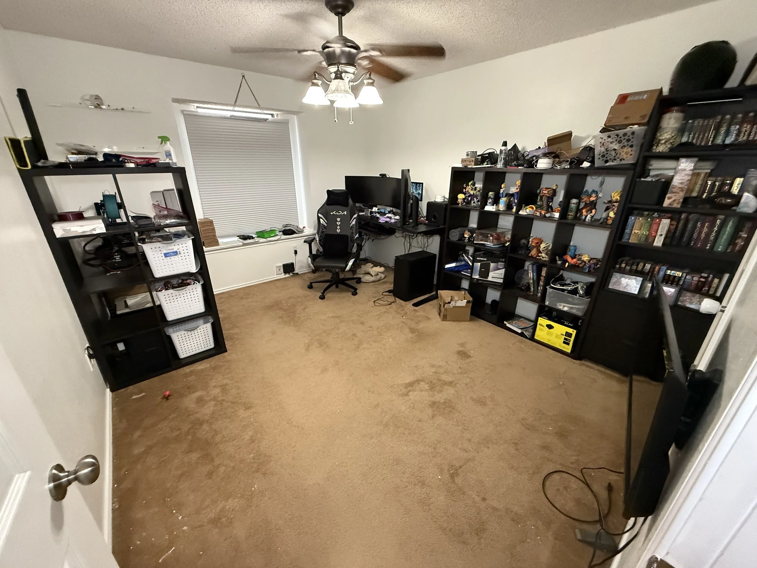 An untidy home office with brown carpet, black shelving filled with various items, a desk with dual monitors, a gaming chair, and a ceiling fan with lights.