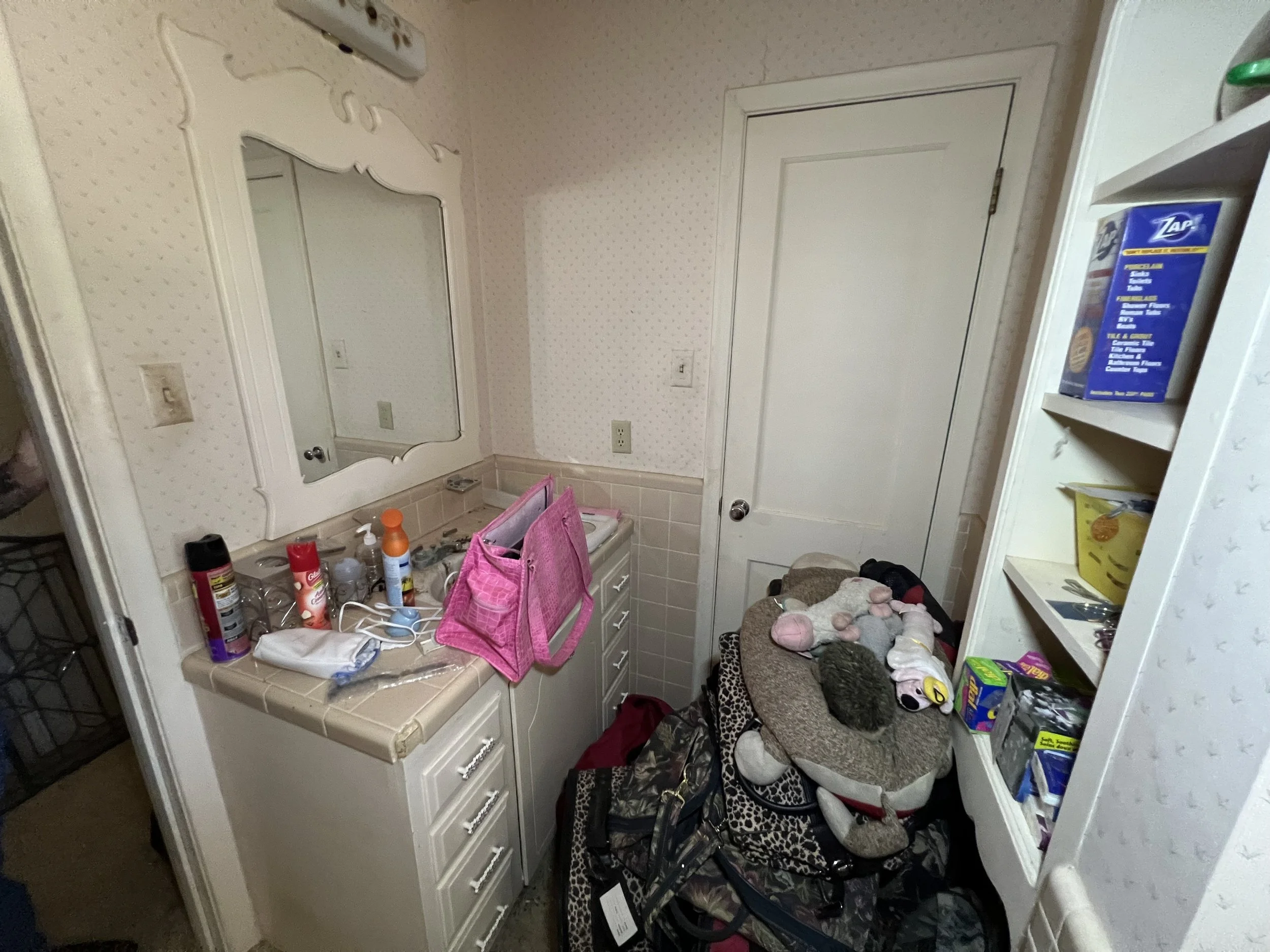 A cluttered bathroom vanity with various toiletries, a pink organizer, and a cluttered corner with bags and stuffed animals.