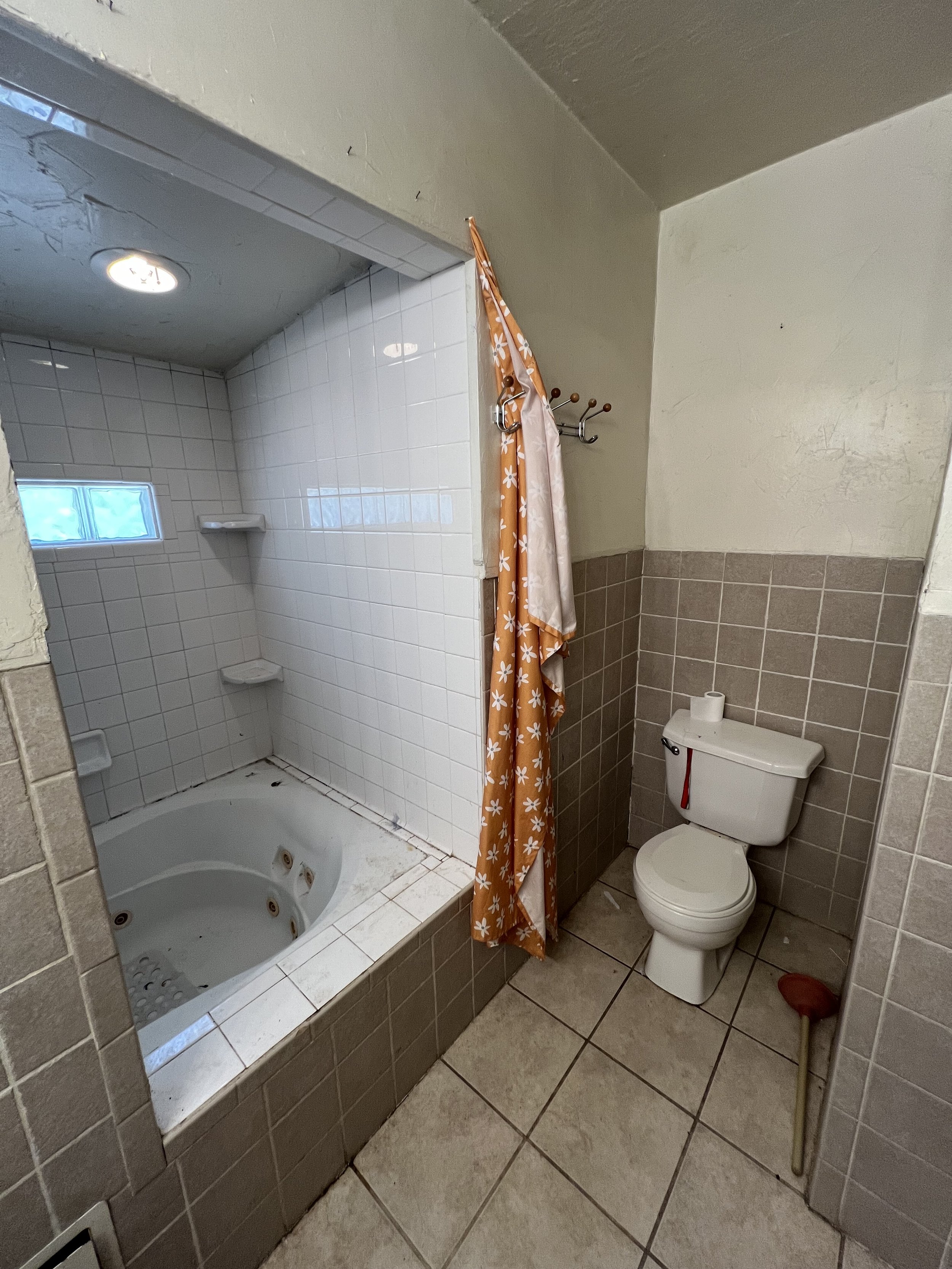 A bathroom with a small shower stall and a toilet, with beige and white tiles on the floor and walls, a small window, and a shower curtain.