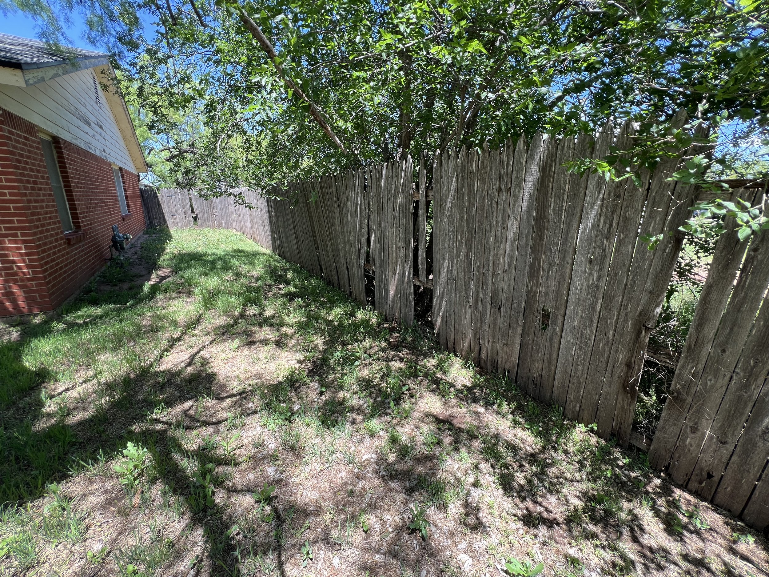 Side yard of a house with a red brick wall, a wooden fence, and some green grass and trees, with shadows cast on the ground.