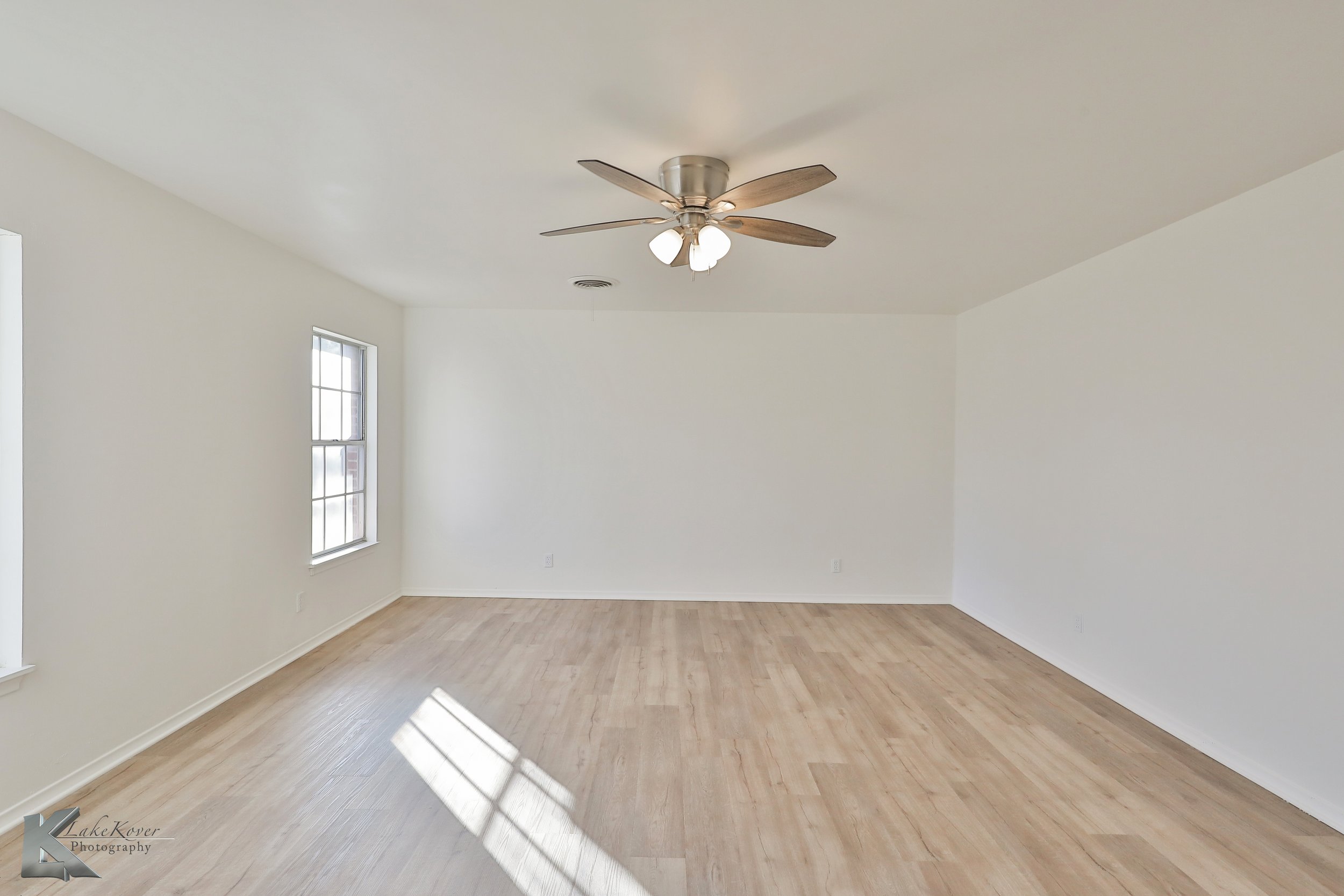 Empty room with wooden floors, white walls, two windows, ceiling fan with lights.
