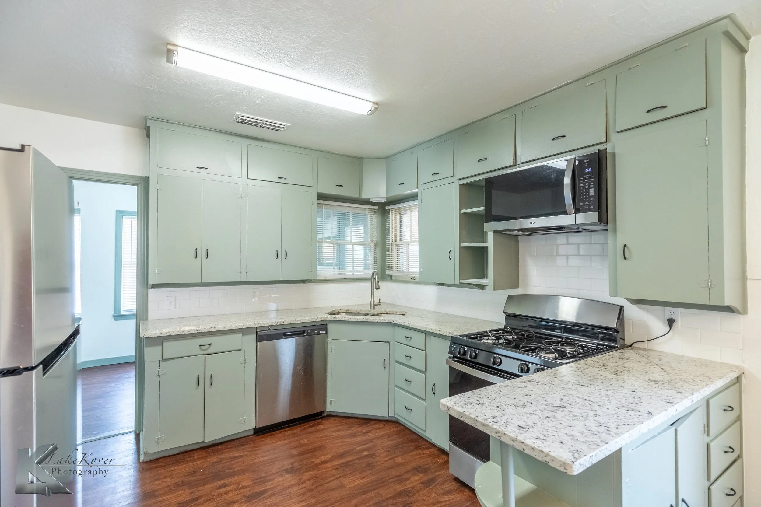 A kitchen with mint green cabinets, granite countertops, stainless steel appliances, a white subway tile backsplash, and hardwood floors.
