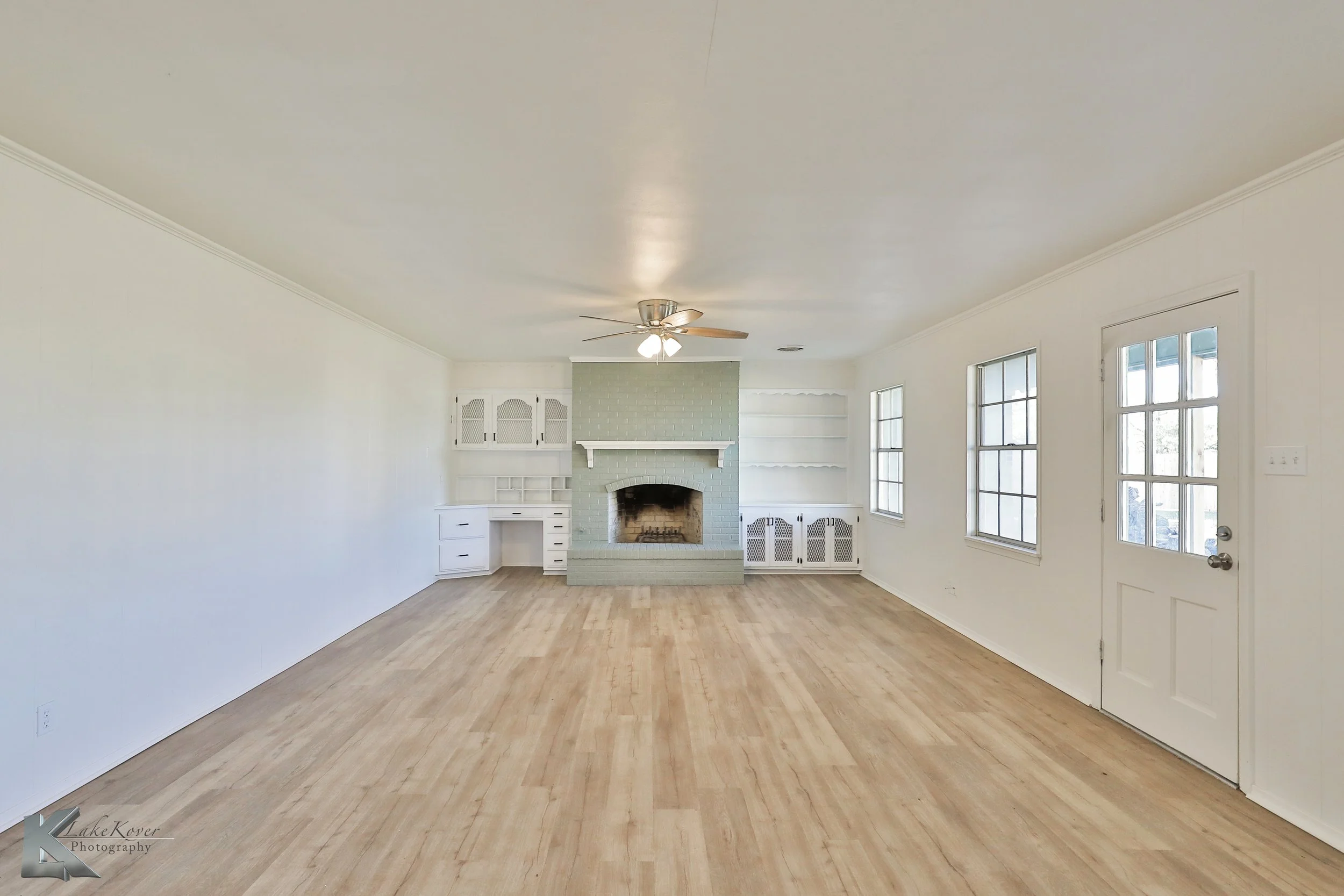 Empty living room with hardwood floors, white walls, multiple windows, a ceiling fan, a fireplace with a white shelf, built-in shelves, and a door leading outside.