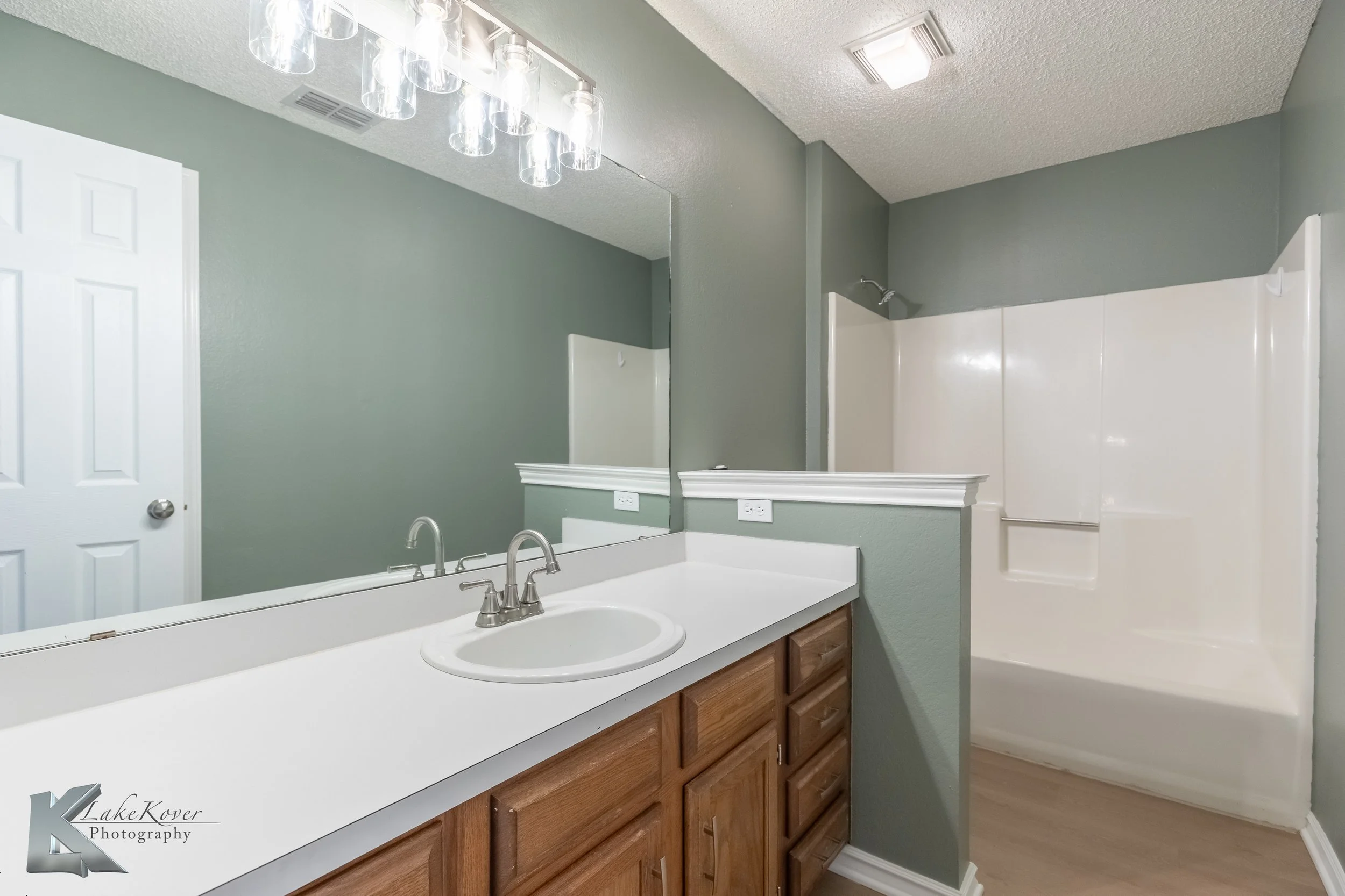 Bathroom with green painted walls, a large mirror above the white sink and wooden cabinets, a shower area with a sliding door, and ceiling lighting.