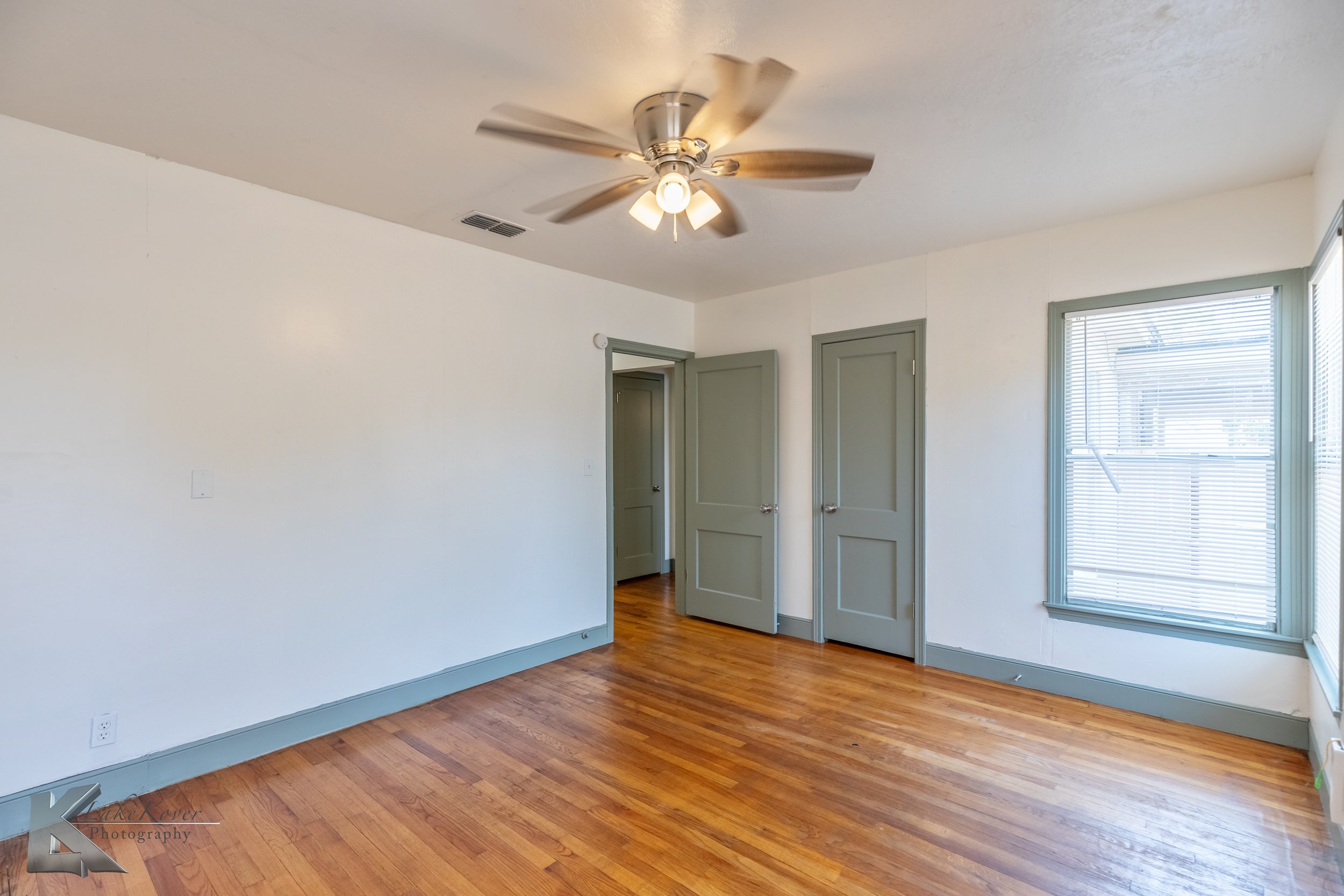 Empty room with hardwood floors, white walls, gray door and window frames, a ceiling fan, and a window with blinds.