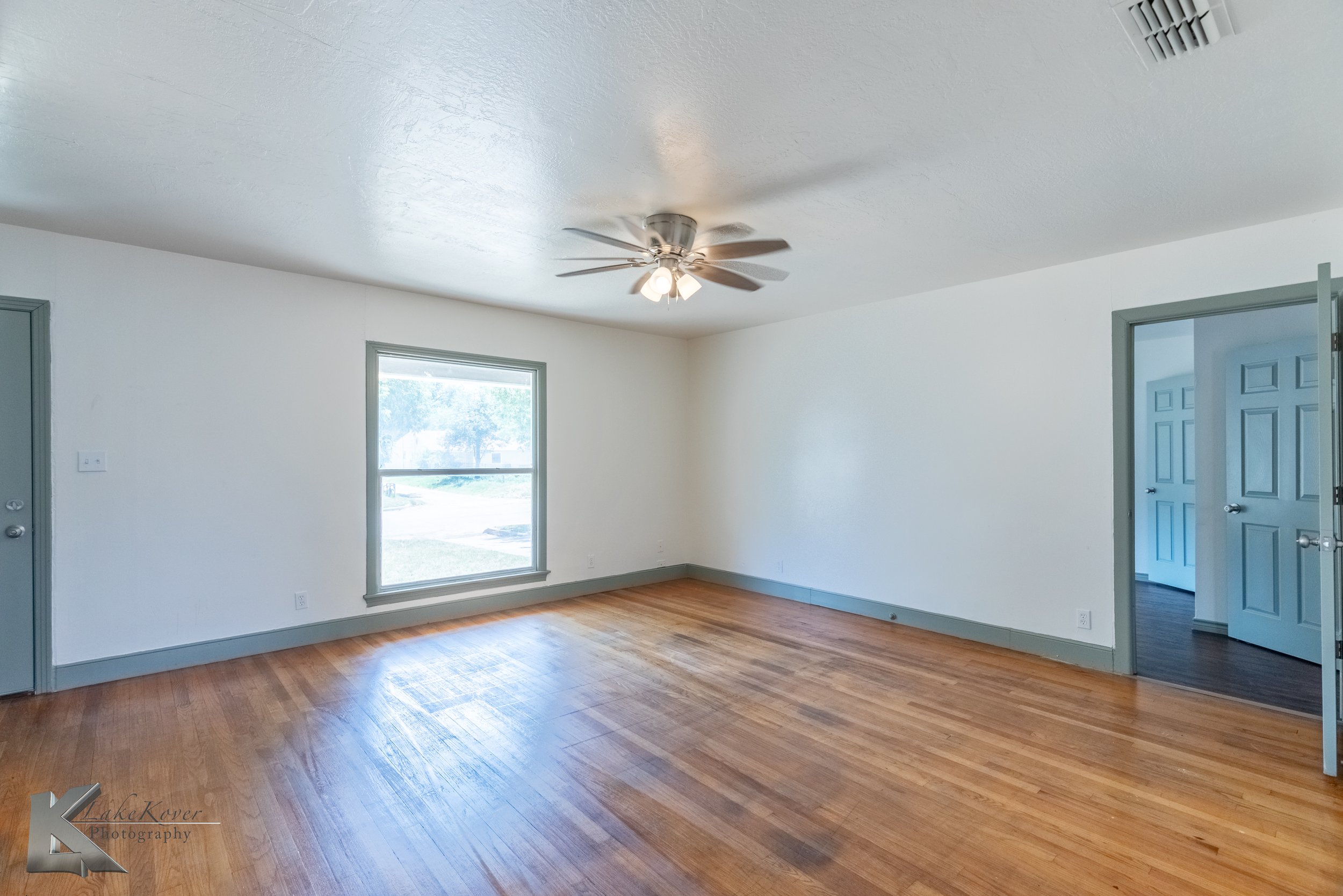 Empty living room with wooden floors, white walls, a ceiling fan with lights, a window, a partially open door, and trim painted in gray.