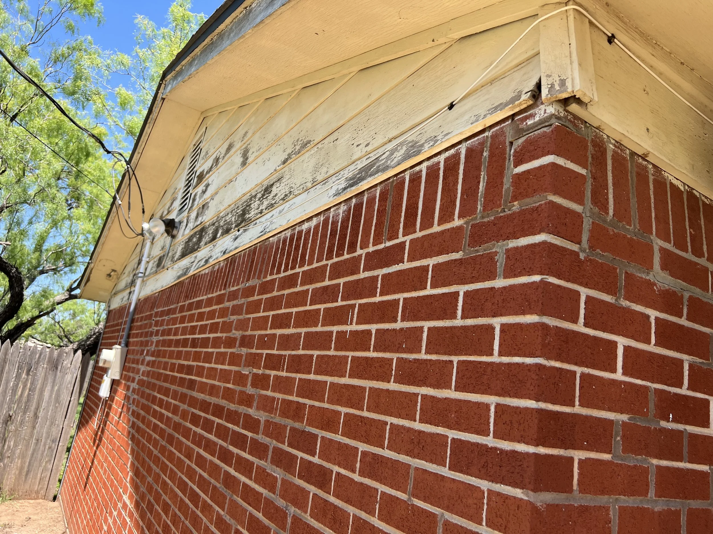 Close-up of a brick wall with a weathered section of siding above it on a house, with some wires and a small light installed, and a tree and blue sky in the background.