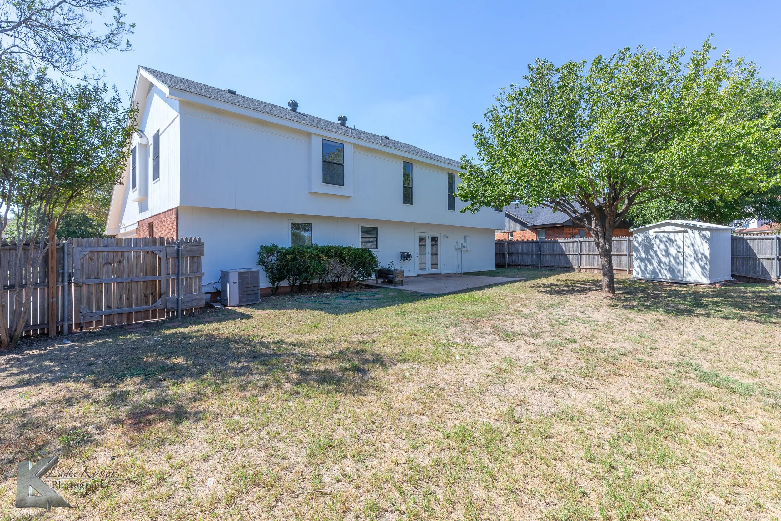 Backyard of a two-story white house with a small concrete patio, surrounded by a wooden fence, with a large tree providing shade and a small white storage shed in the corner.