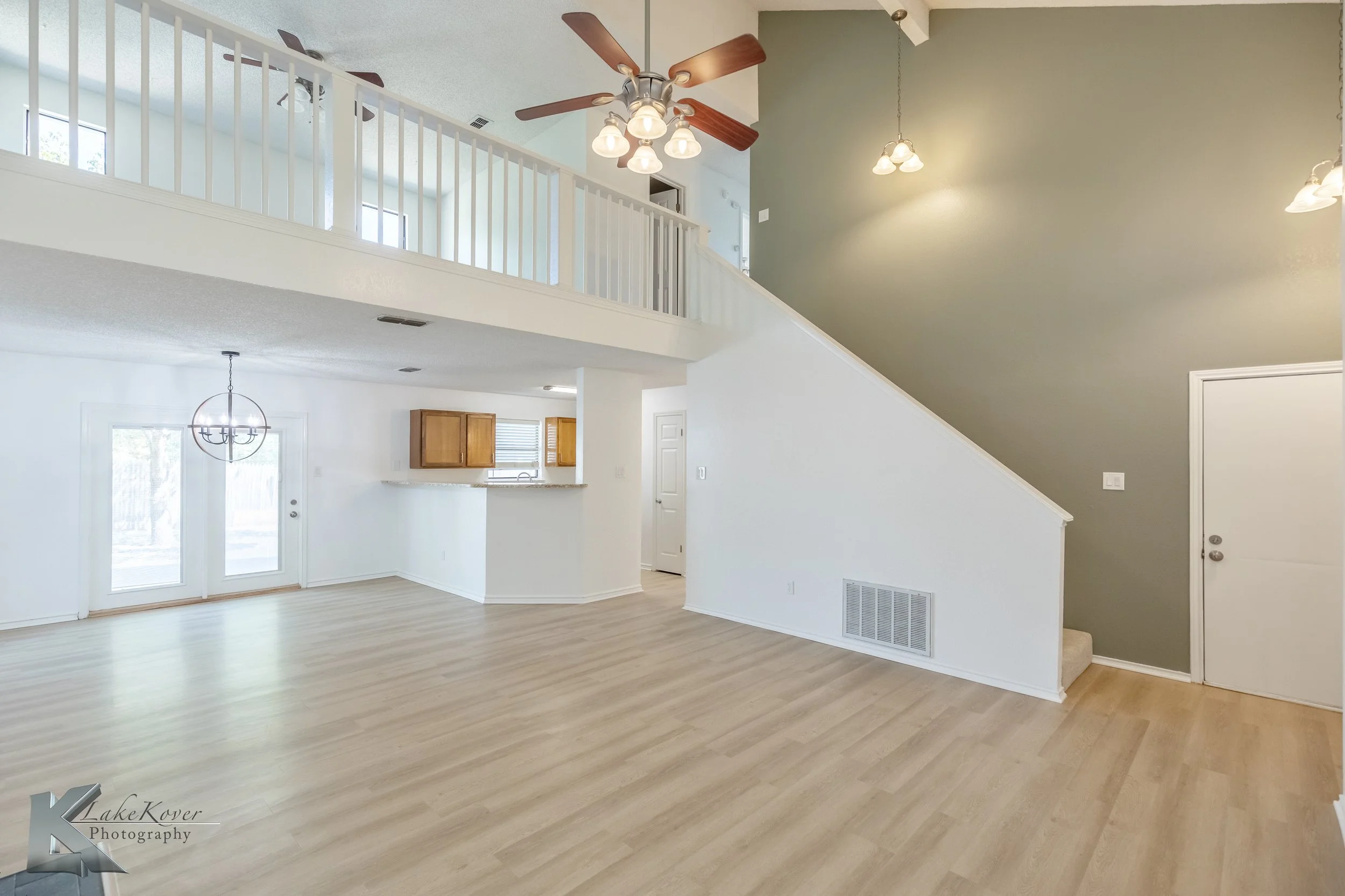 Interior of a bright, open living room with light wood floors, white walls, and a staircase with a green accent wall, ceiling fan, and multiple hanging lights. The back wall features double glass doors leading outside, and there is access to a kitche